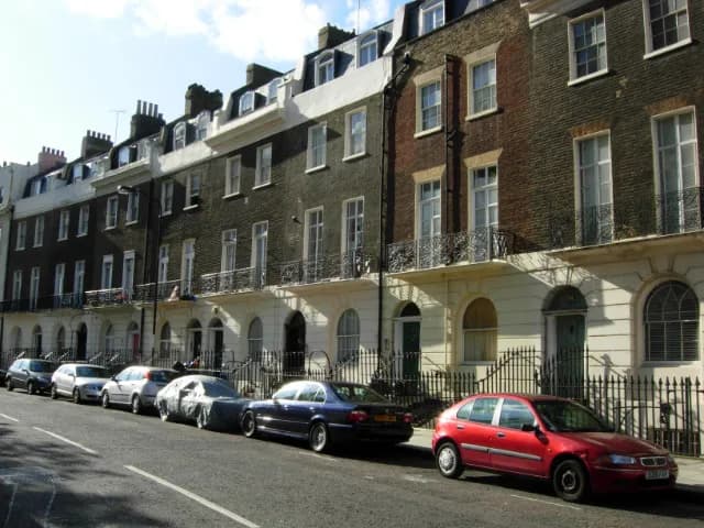 A row of brick apartment buildings with white trim and balconies, with cars parked along the street in front of them.