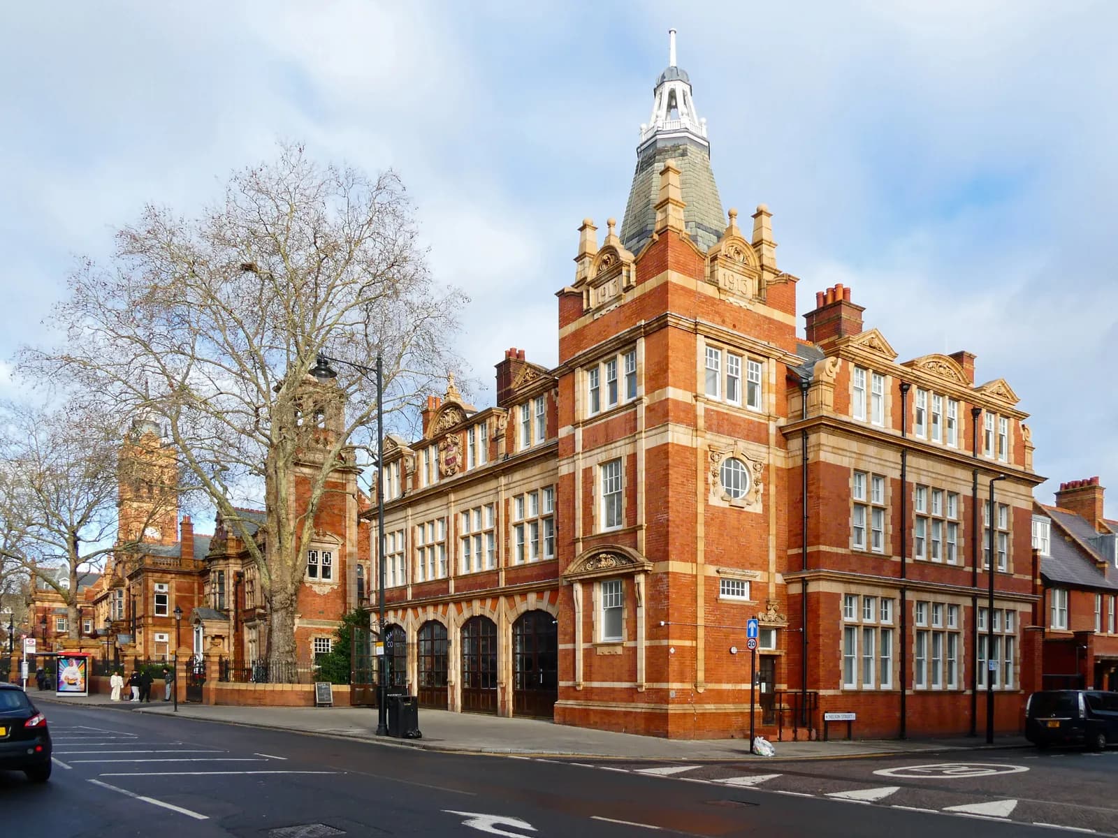 A large red brick building with a tall spire on the right, and other similar buildings and a large tree further down the street on the left.