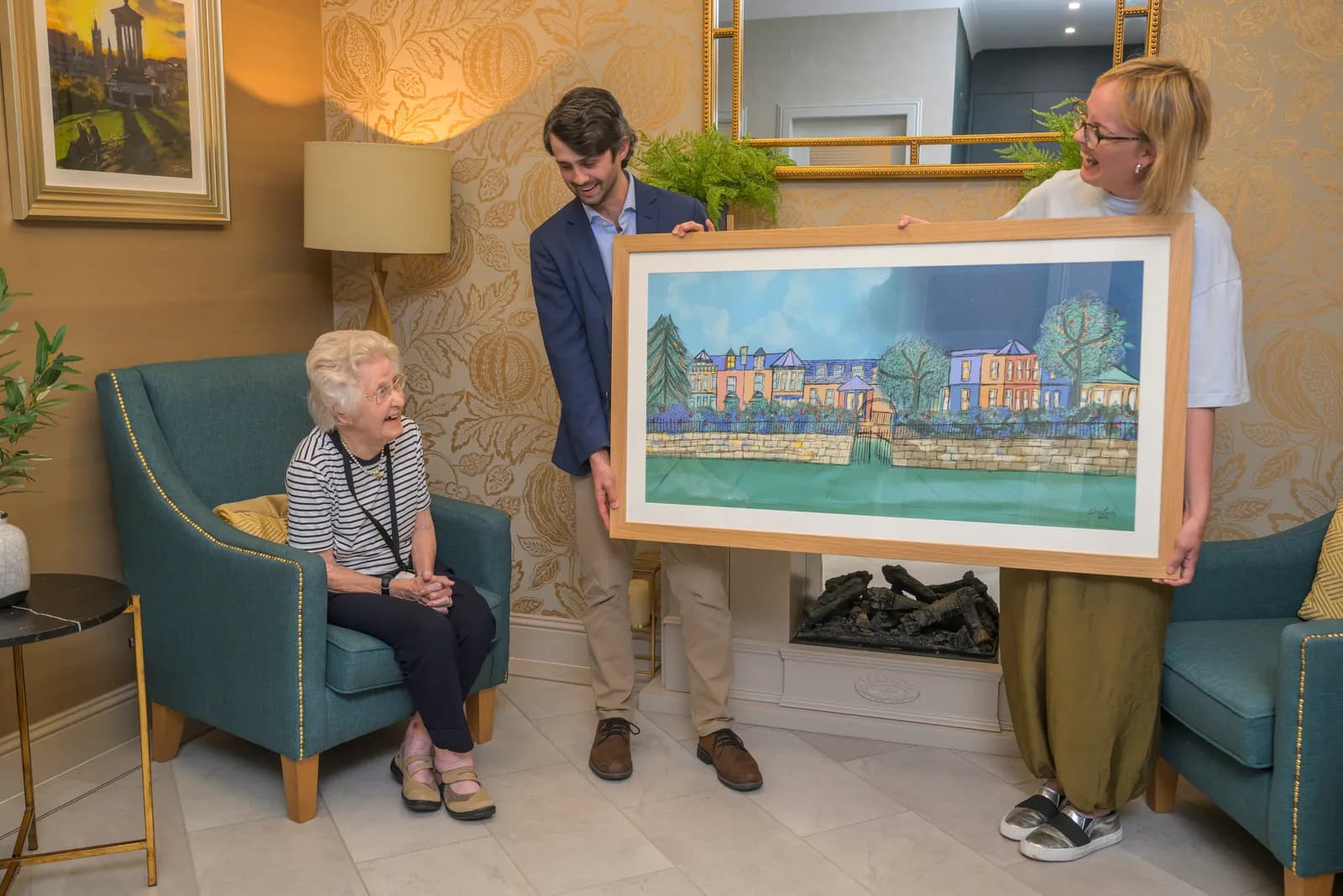 An elderly woman seated in a teal armchair smiles as a man and a woman hold up a framed painting of a waterside town.
