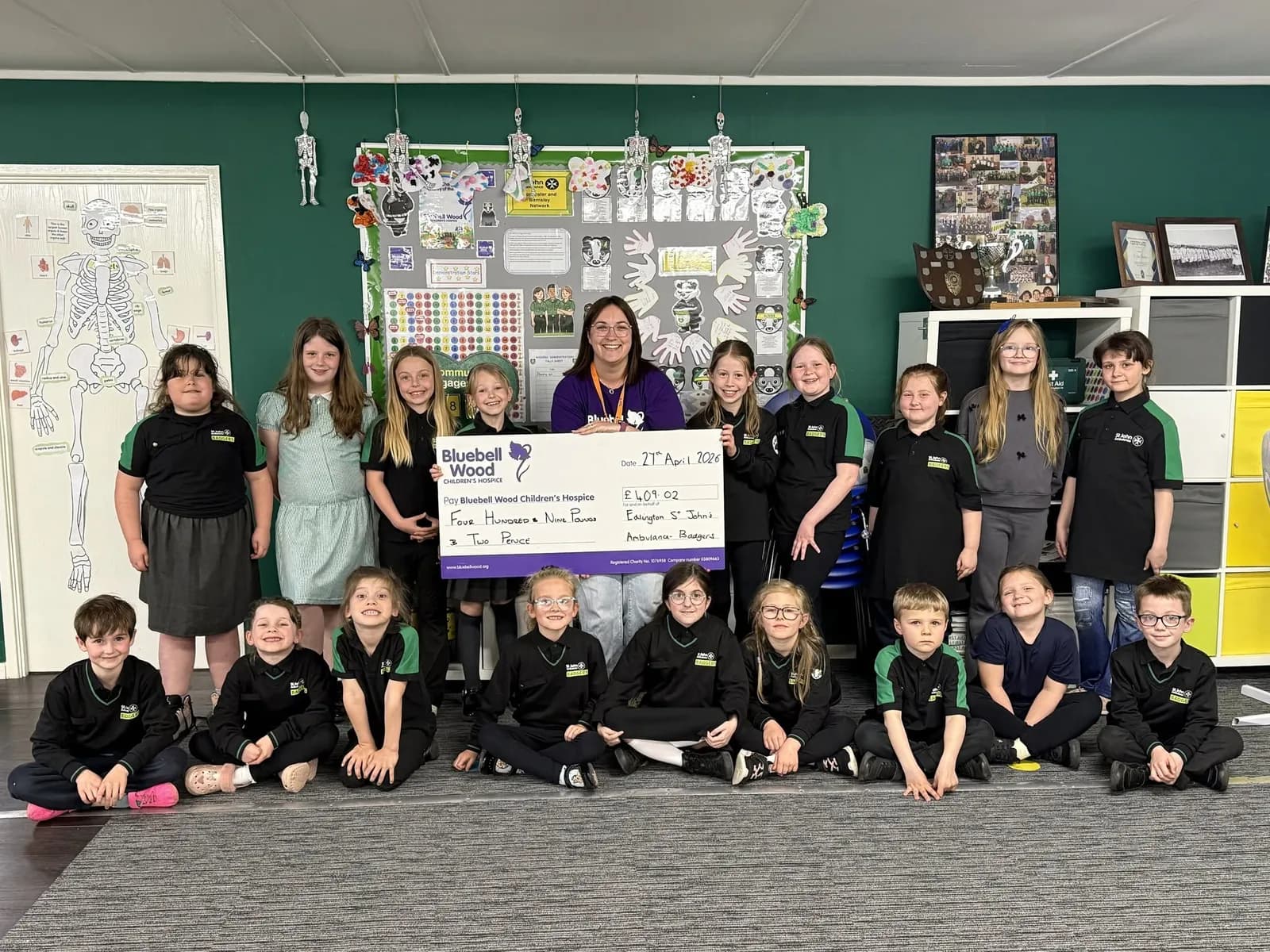 A group of children and an adult woman gather in a classroom holding up a large check made out to Bluebell Wood Children's Hospice.