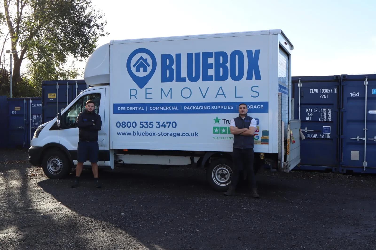 Two men standing confidently in front of a white Bluebox Removals truck, with blue shipping containers in the background.