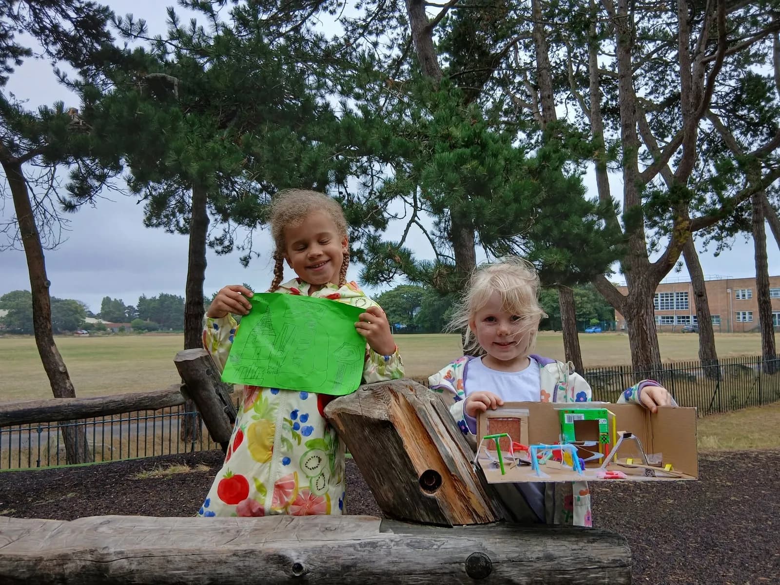 Two young girls in raincoats display a green drawing and a cardboard model of a playground, standing in front of a field with trees and a building in the background.