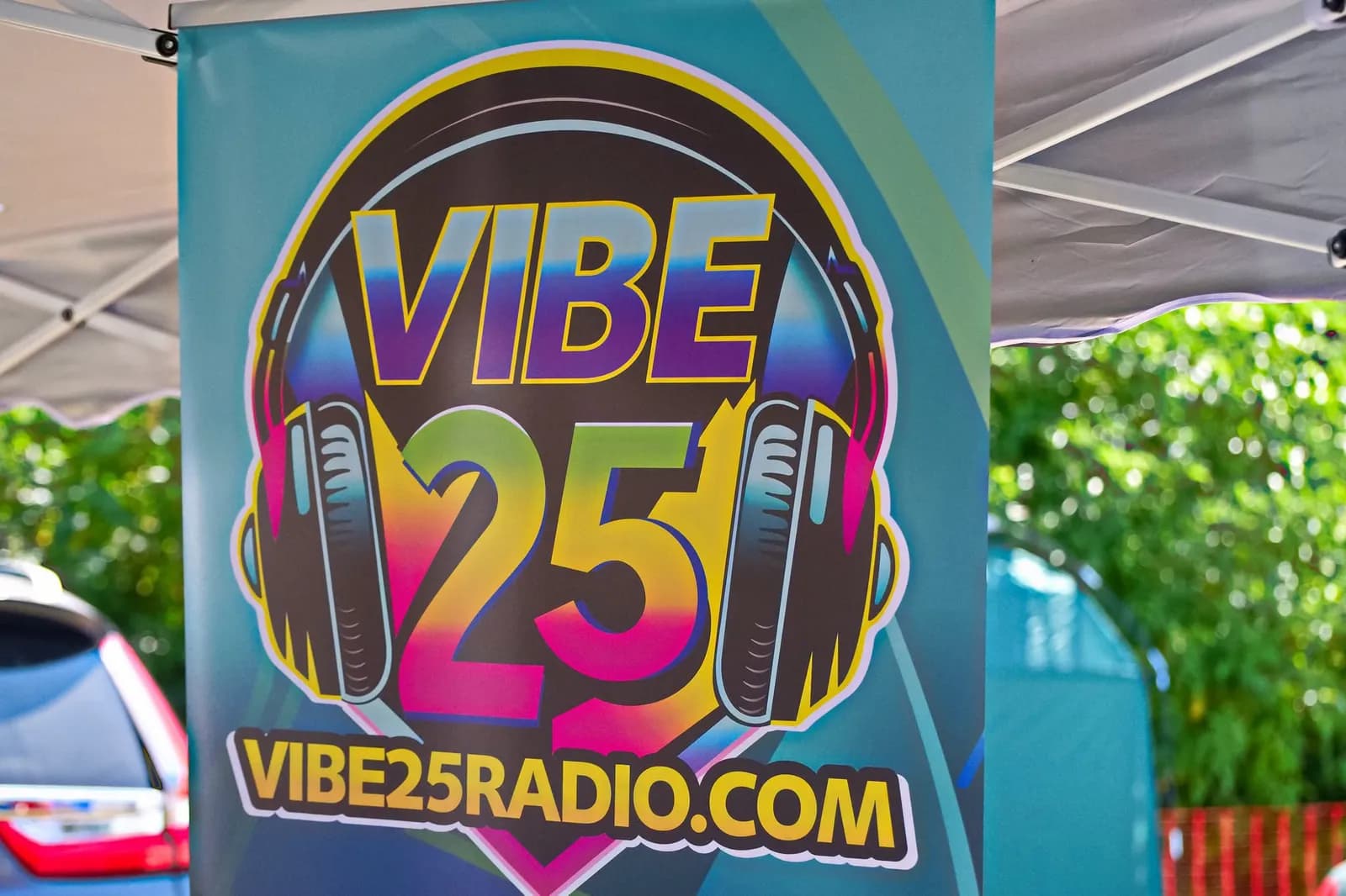 A VIBE 25 RADIO banner with headphones and a website address is displayed under a white tent, with blurred green trees in the background.