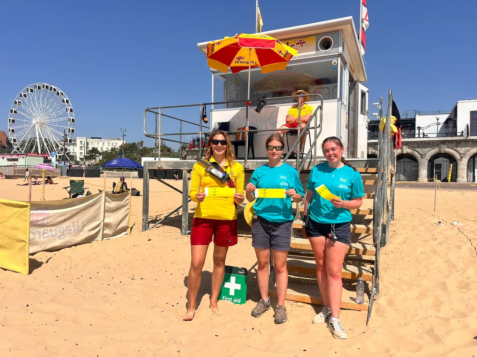 Three young women stand on a sandy beach next to a lifeguard tower, holding yellow slips of paper, with a Ferris wheel and buildings visible in the background.