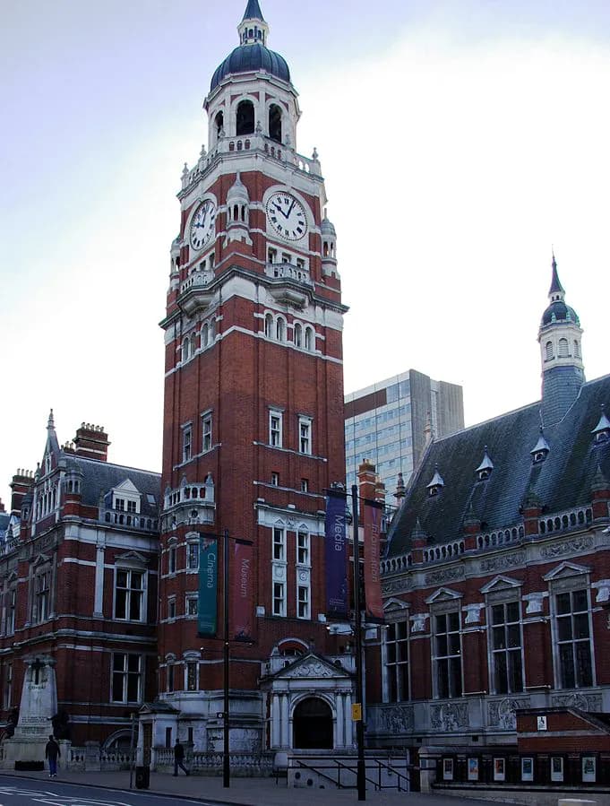 A towering red brick clock tower with white accents and a dark domed roof stands prominently against a light sky, flanked by other red brick buildings with detailed architecture and banners.