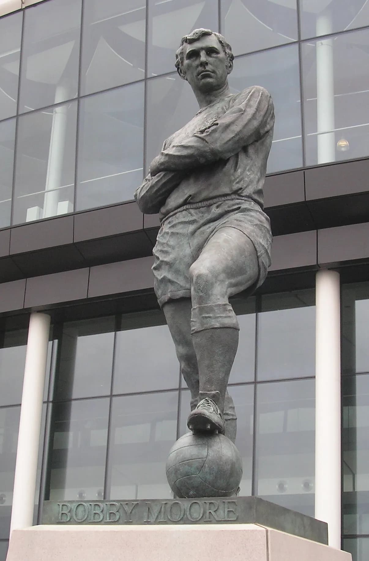 A bronze statue of Bobby Moore in a soccer uniform with his arms crossed, one foot resting on a soccer ball, in front of a modern glass building.