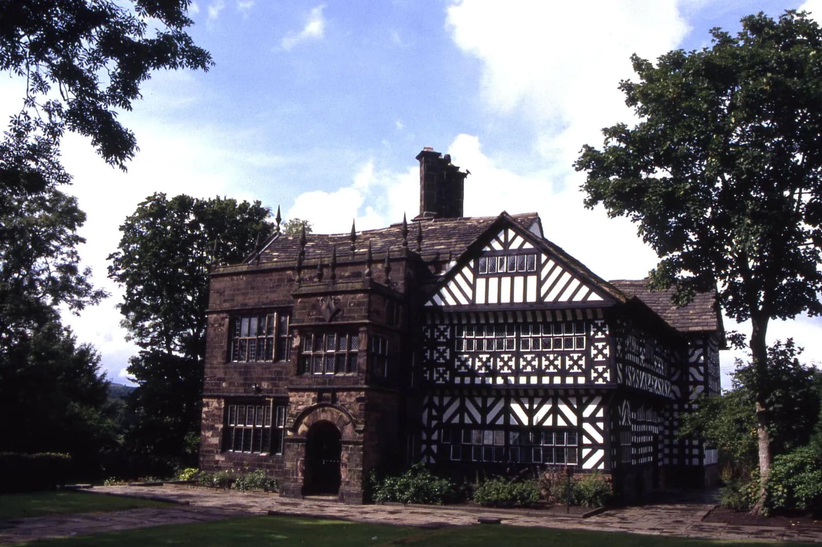 A historic, dark stone and half-timbered building with multiple windows stands prominently amidst green trees and a grassy field under a bright sky with scattered clouds.
