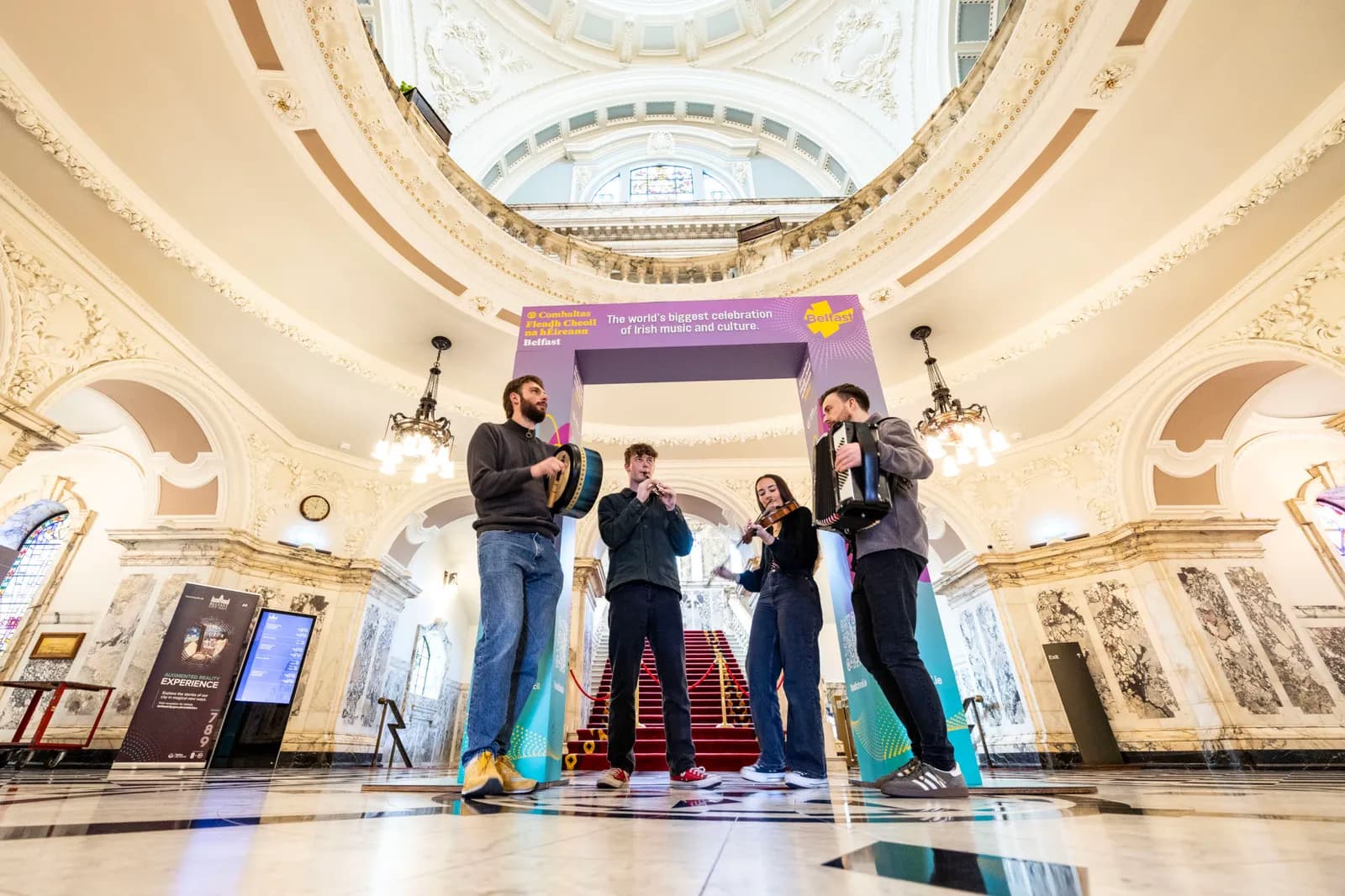 Four musicians, two men and two women, dressed in casual attire, play Irish instruments under a banner for an Irish music and culture festival inside a grand hall with ornate architecture.