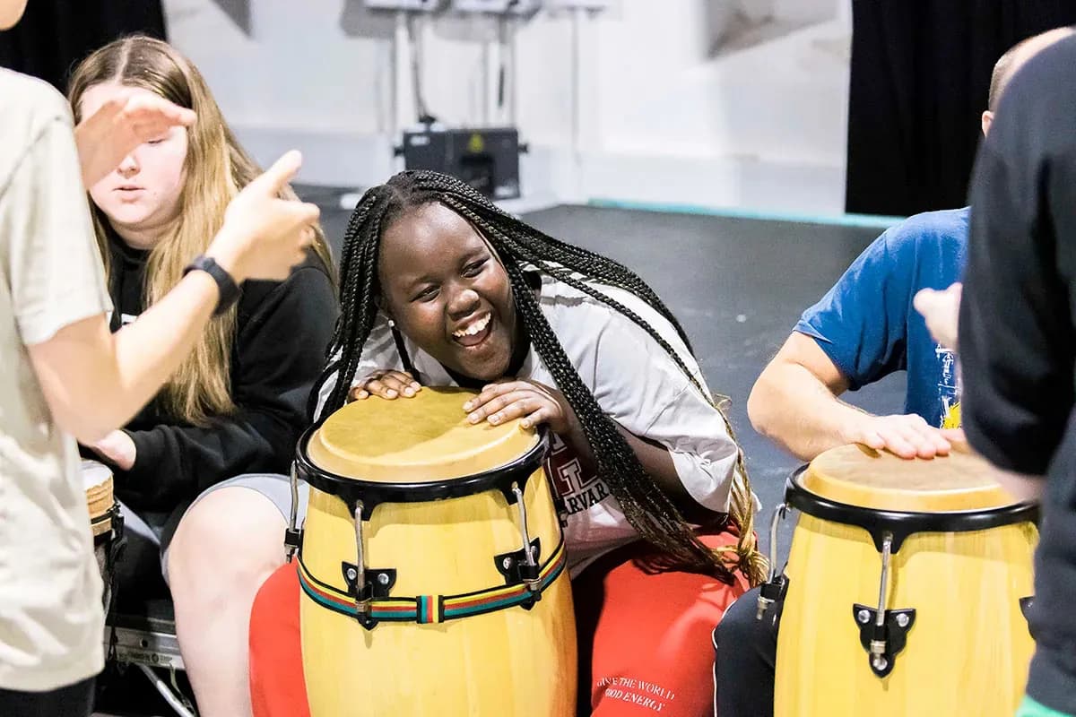 A young Black woman with braided hair smiles broadly while playing a yellow conga drum with others around her.