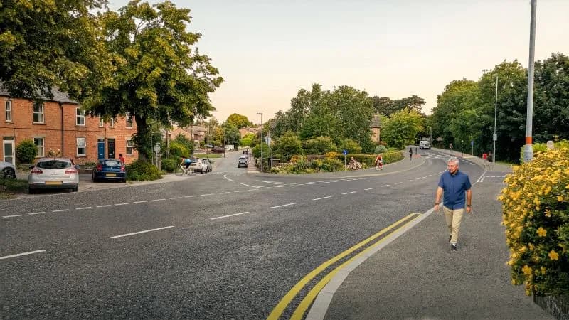 A man in a blue shirt and khaki pants walks down a path next to a road, with brick buildings and trees lining the street.