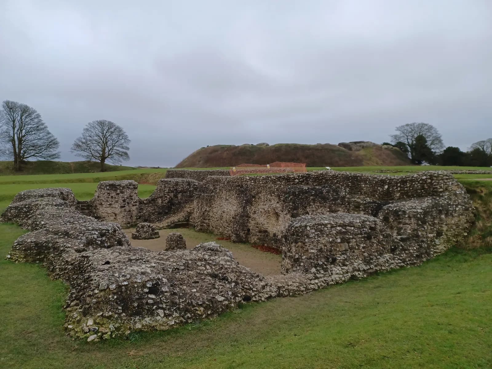 Ancient Roman ruins with low stone walls stand on a green lawn beneath a cloudy sky, with distant trees and an earthen mound in the background.