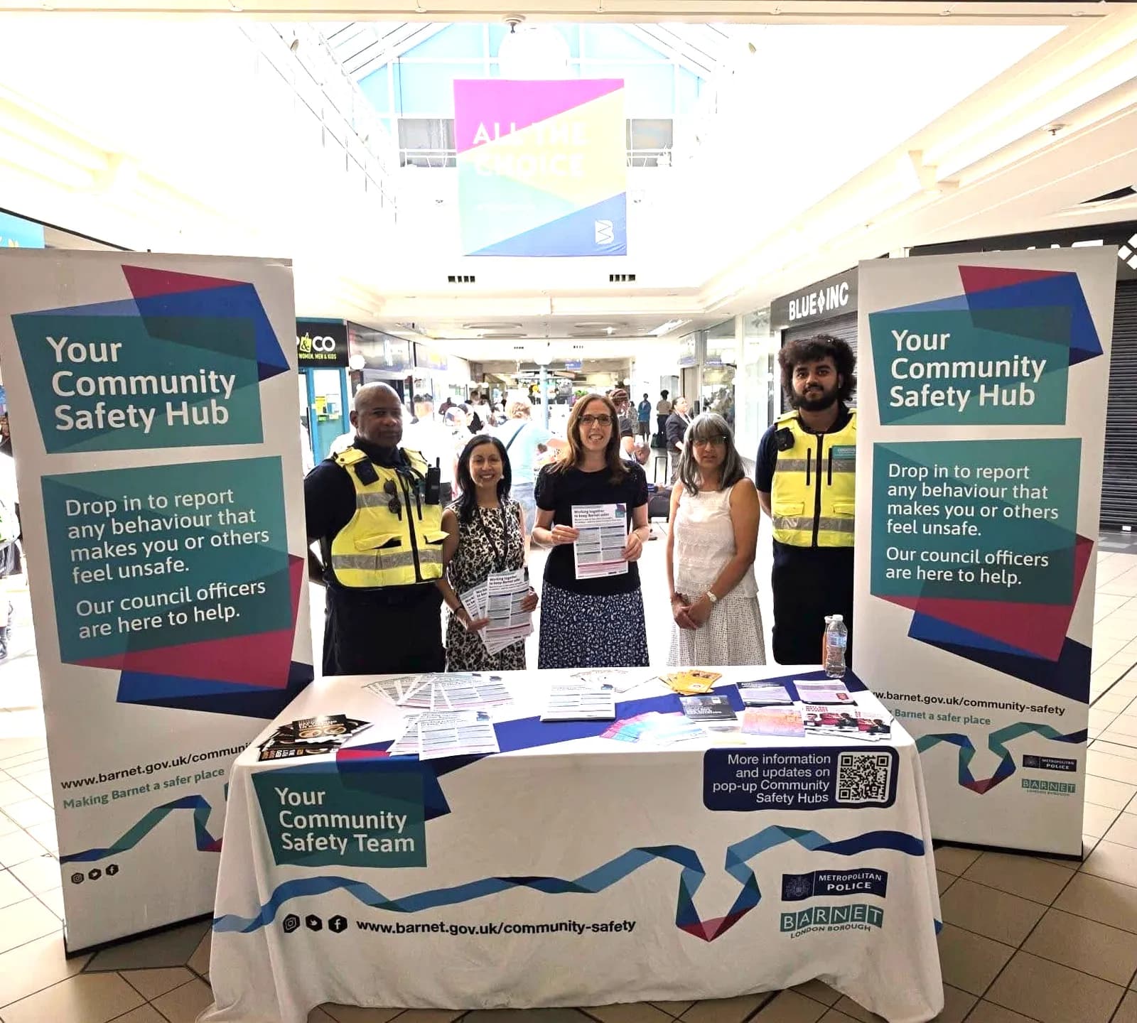 Five community safety officers stand behind a table with informational fliers under a sign that reads, "Your Community Safety Hub".