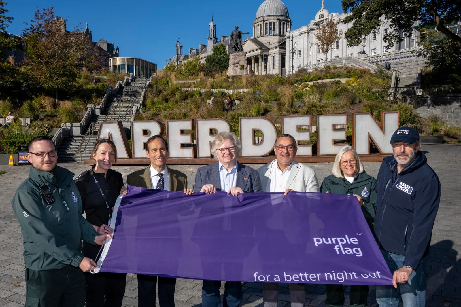 Seven people stand in front of large white letters spelling 'ABERDEEN' on a hillside, holding a purple flag that reads 'purple flag for a better night out'.