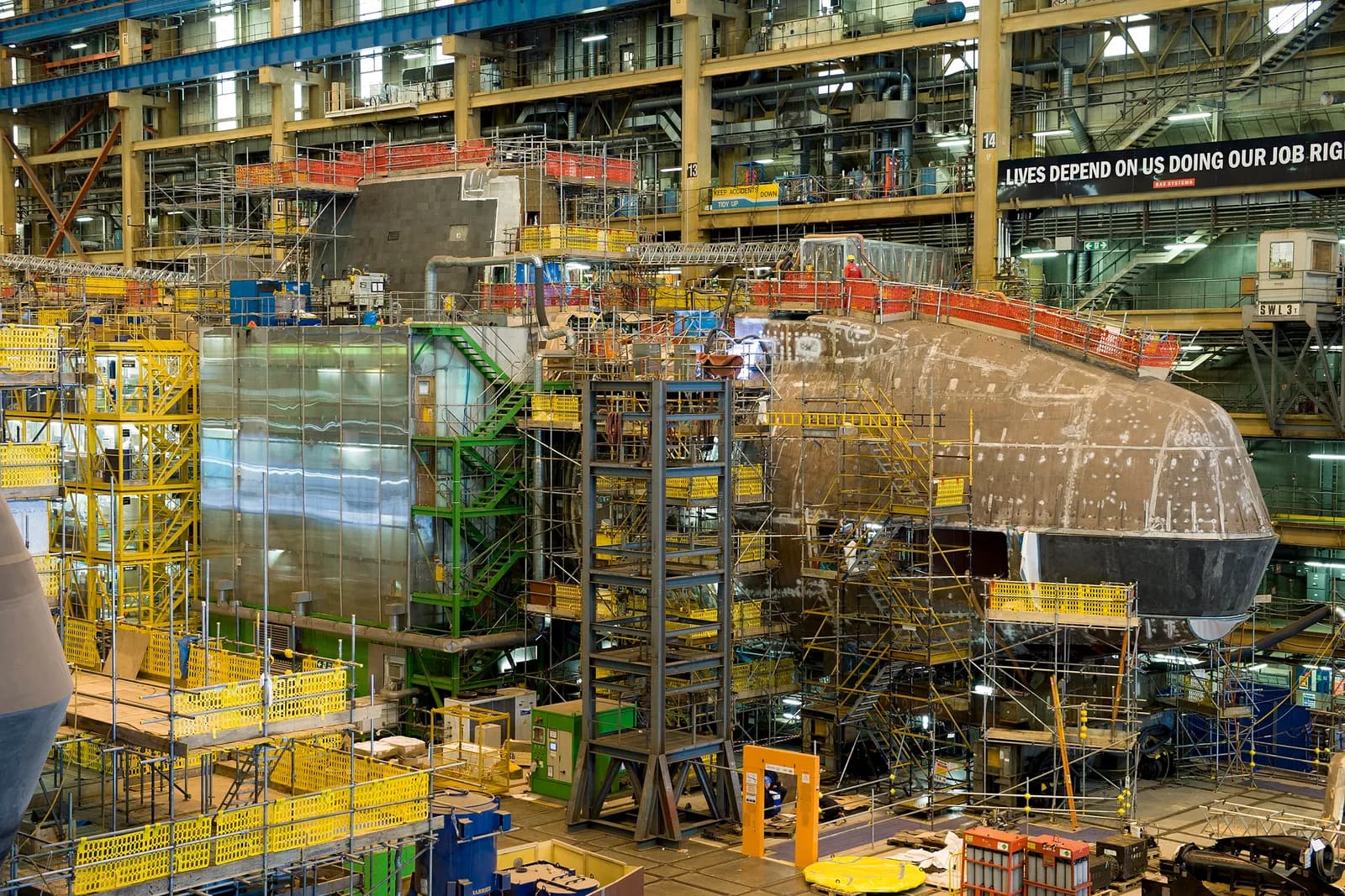 A large, unfinished submarine hull surrounded by extensive scaffolding and industrial equipment inside a spacious factory building with a motivational banner reading "LIVES DEPEND ON US DOING OUR JOB RIG..." in the background.