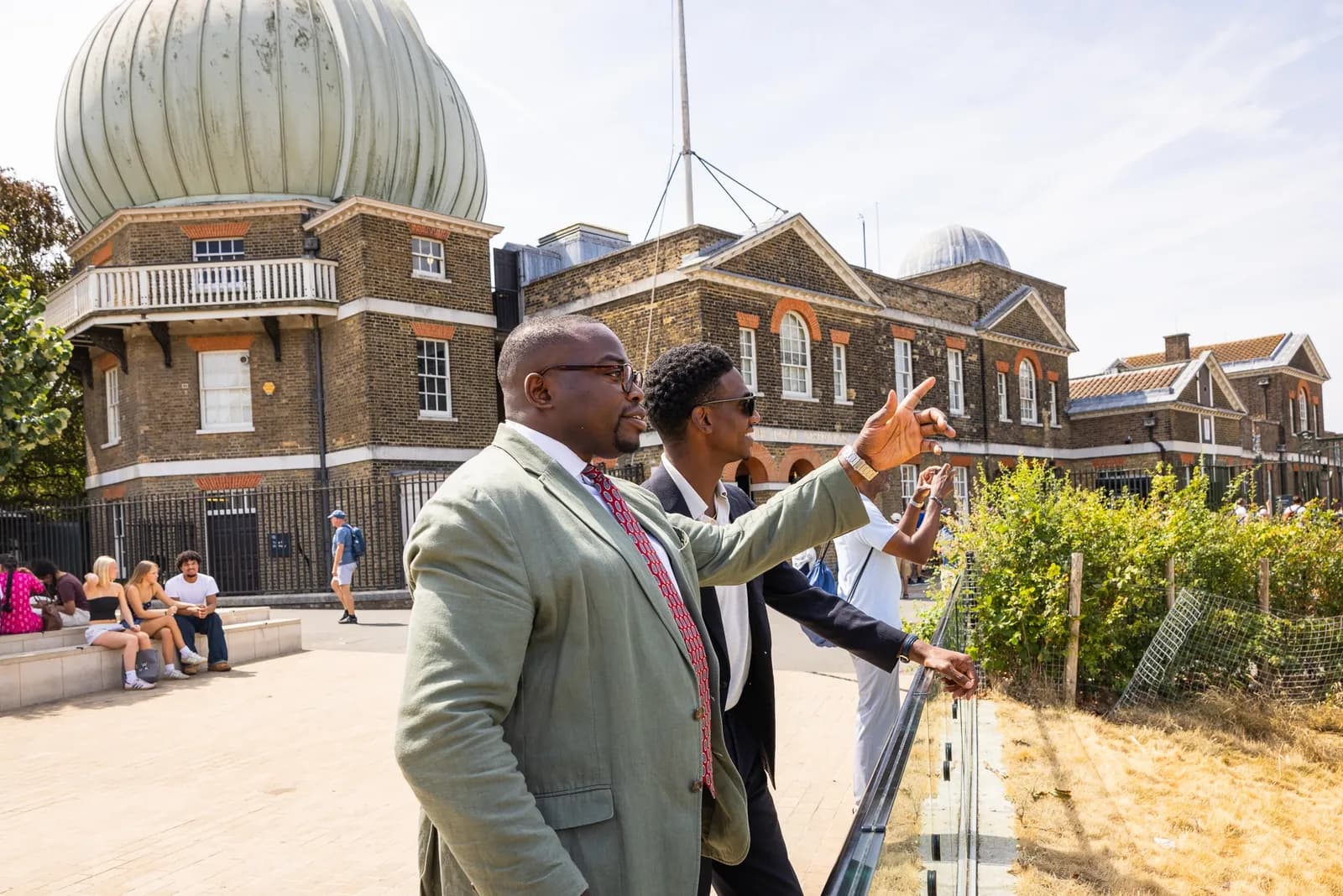 Two men on a bright day stand outside, with one man in a light green suit pointing towards a historic building with a domed roof in the background.
