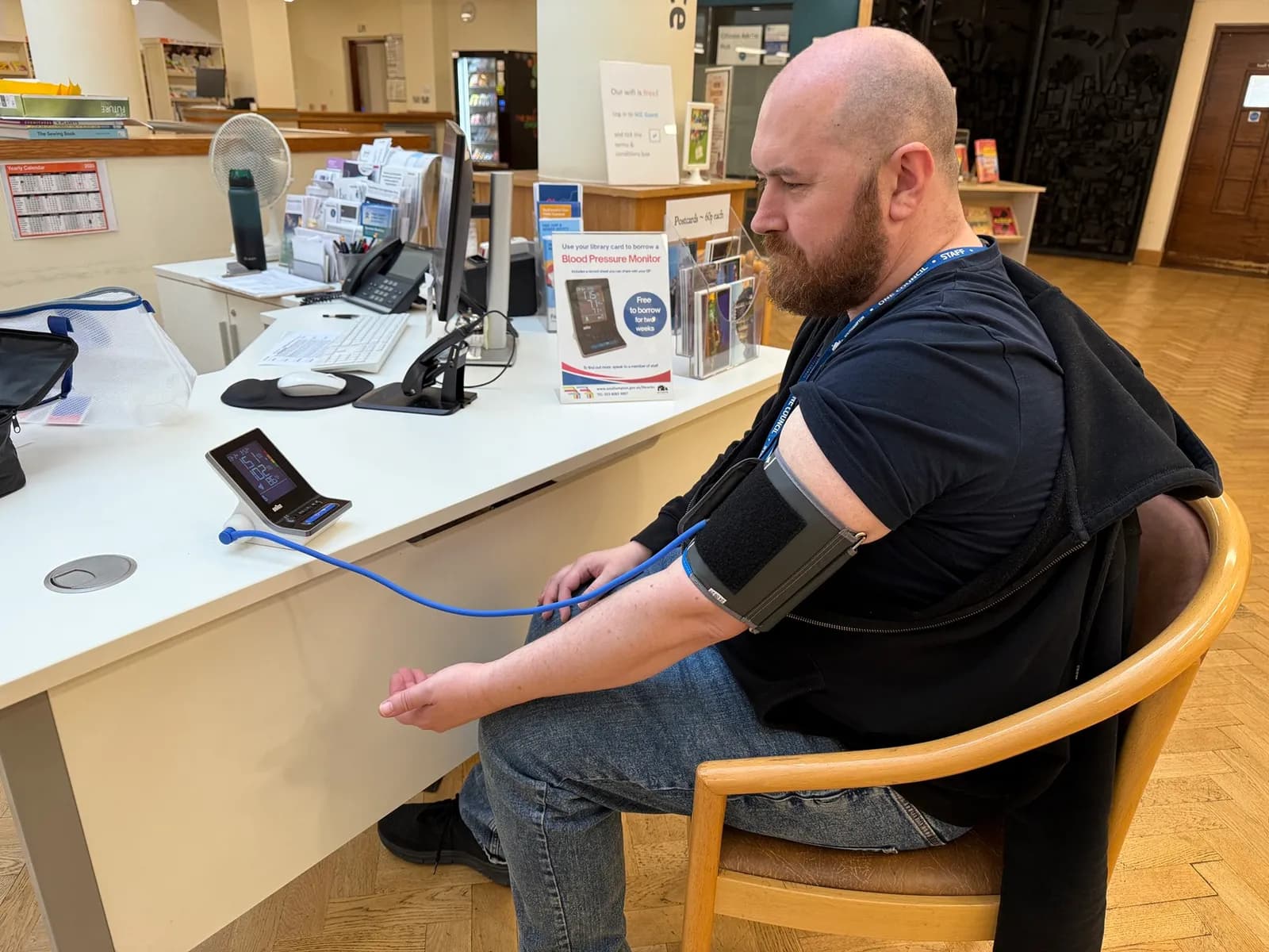 A man with a beard sits in a wooden chair with a blood pressure cuff on his left arm, which is connected by a blue tube to a digital blood pressure monitor on a white desk.