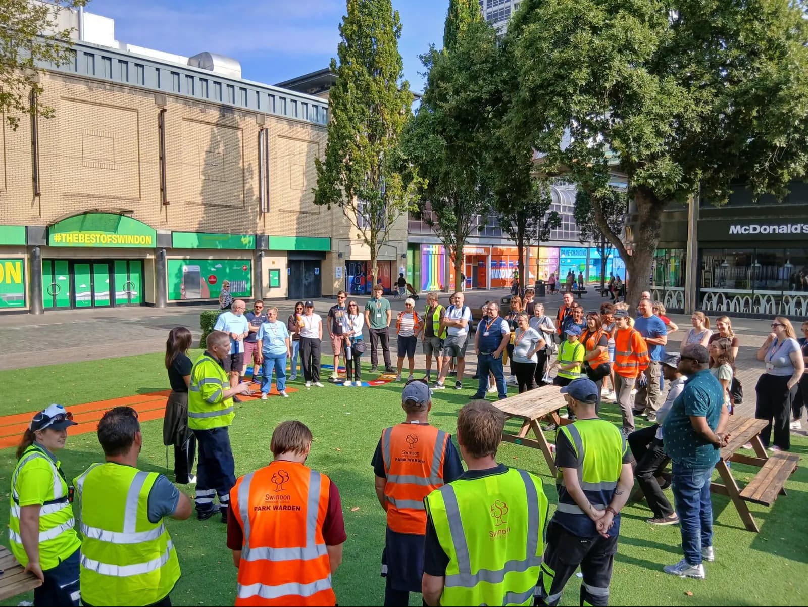 A group of people wearing bright vests are gathered in a circle on a patch of artificial grass in a town square, with buildings and trees in the background.