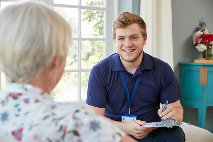 A smiling young man in a blue polo shirt and lanyard is writing on a clipboard while talking to an elderly woman whose back is to the camera.