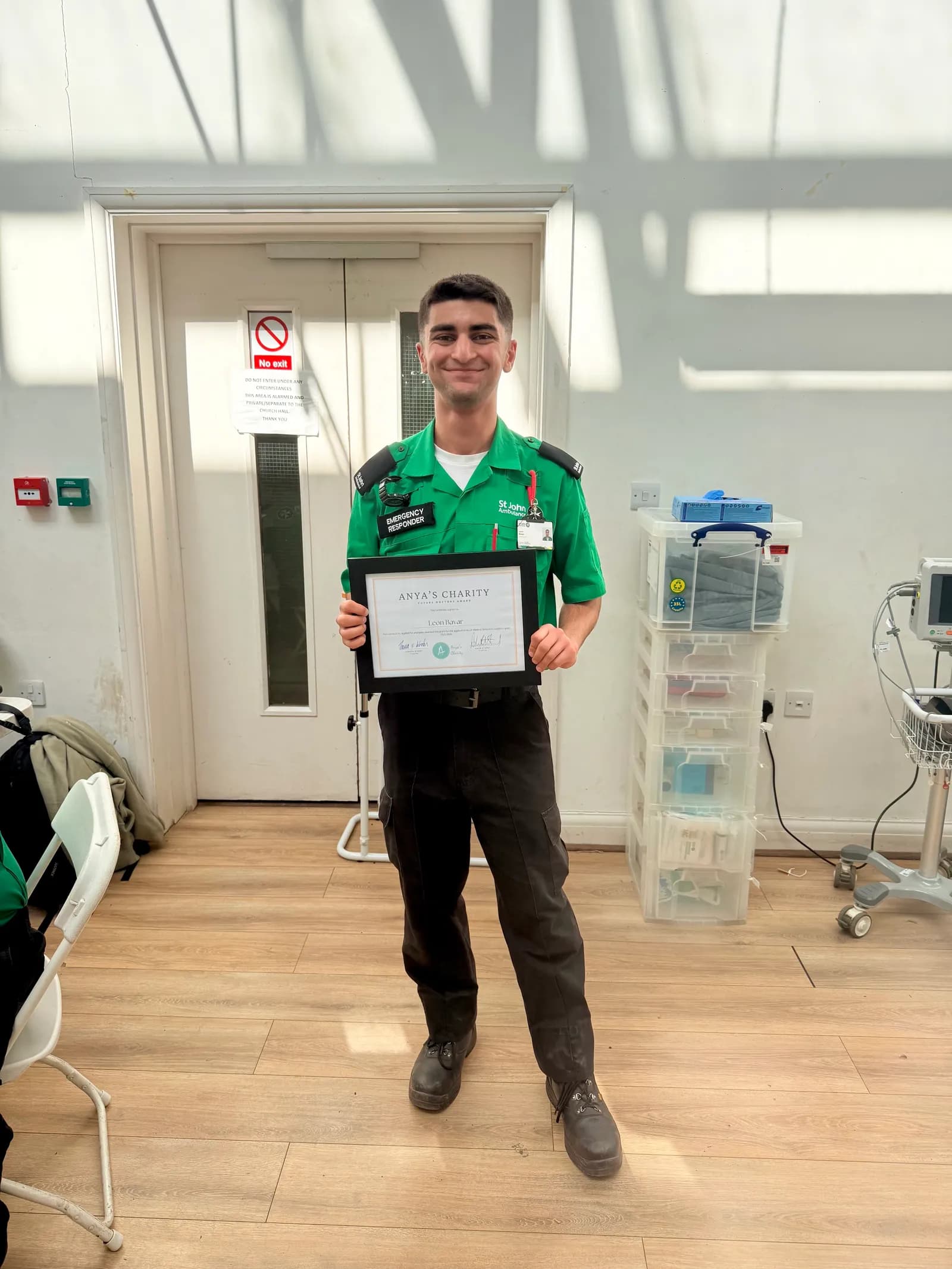 A young man in a green St. John Ambulance uniform smiles while holding a framed certificate for Anya's Charity.