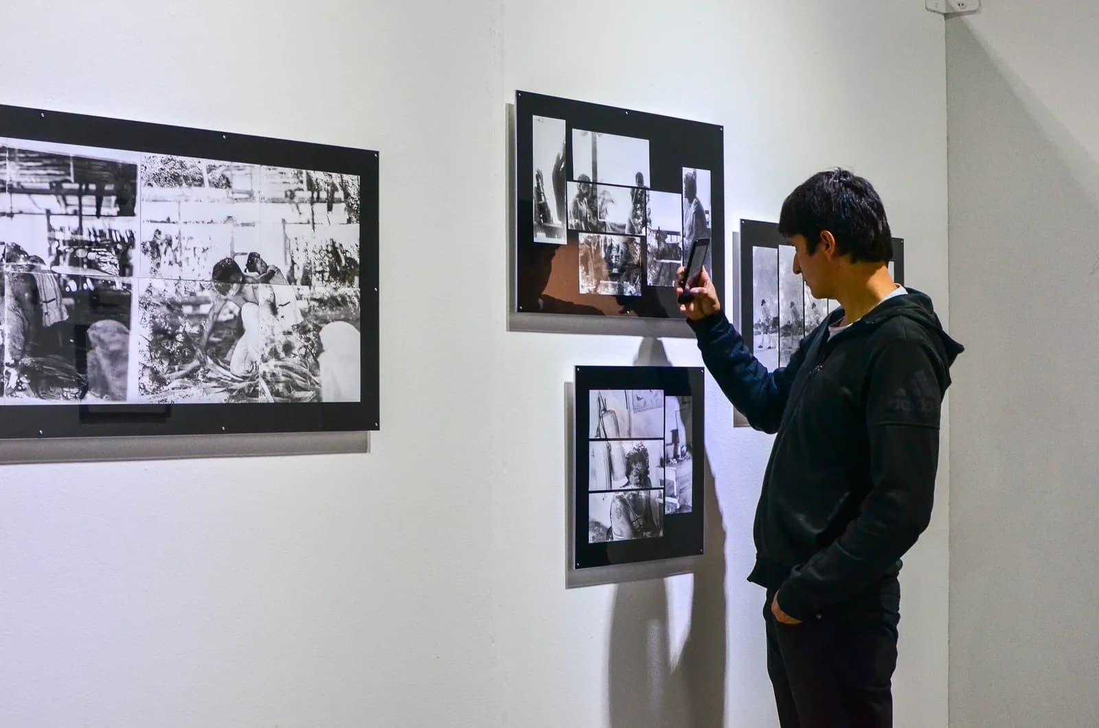 A person in a black hoodie holds up their phone to take a picture of a black and white photo collage on a gallery wall.