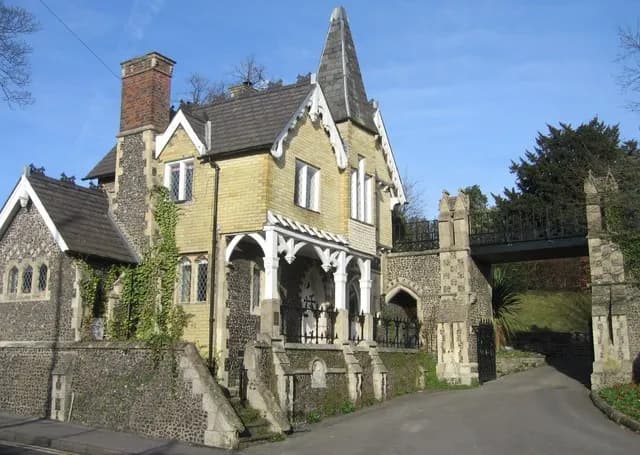 A yellow-bricked house with a tall, pointed spire and white trim is next to a stone archway with a heavy, black gate and a bridge overhead.