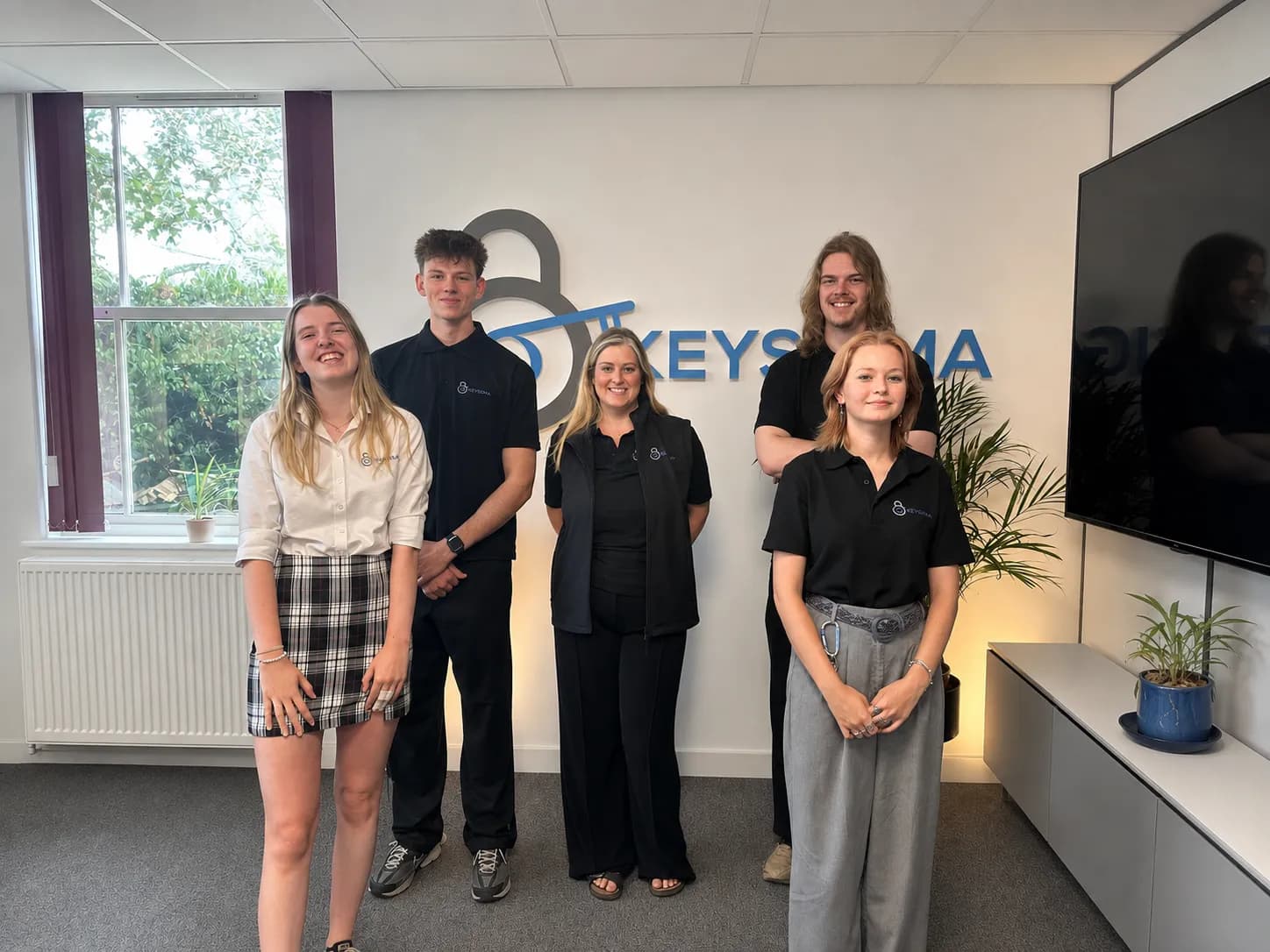 Five young people standing in an office with a company logo on the wall behind them.