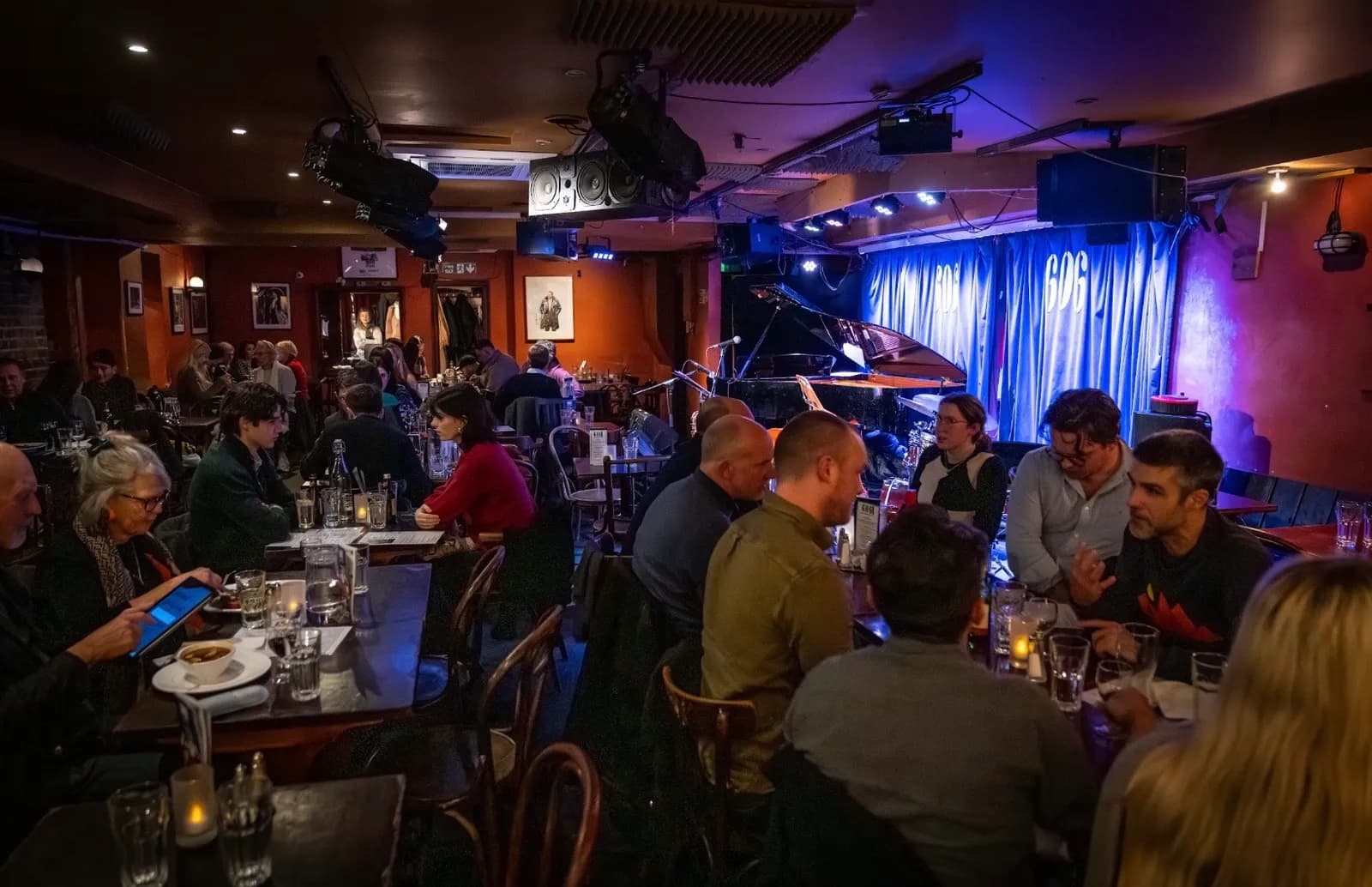 Patrons sit at tables inside a dimly lit jazz club with a piano on a stage in the background.