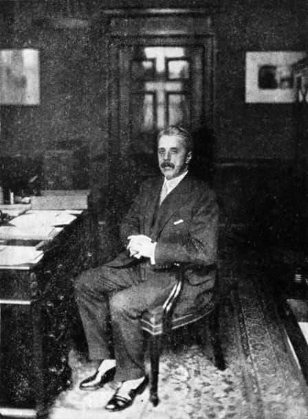 A black and white photograph shows a man with a mustache in a suit sitting at a desk with his hands clasped.