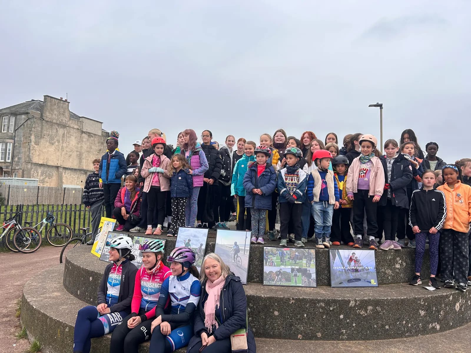 A large group of children and a few adults pose for a picture outdoors, some holding framed photos of bicycles and cyclists, with old buildings and bicycles in the background under an overcast sky.