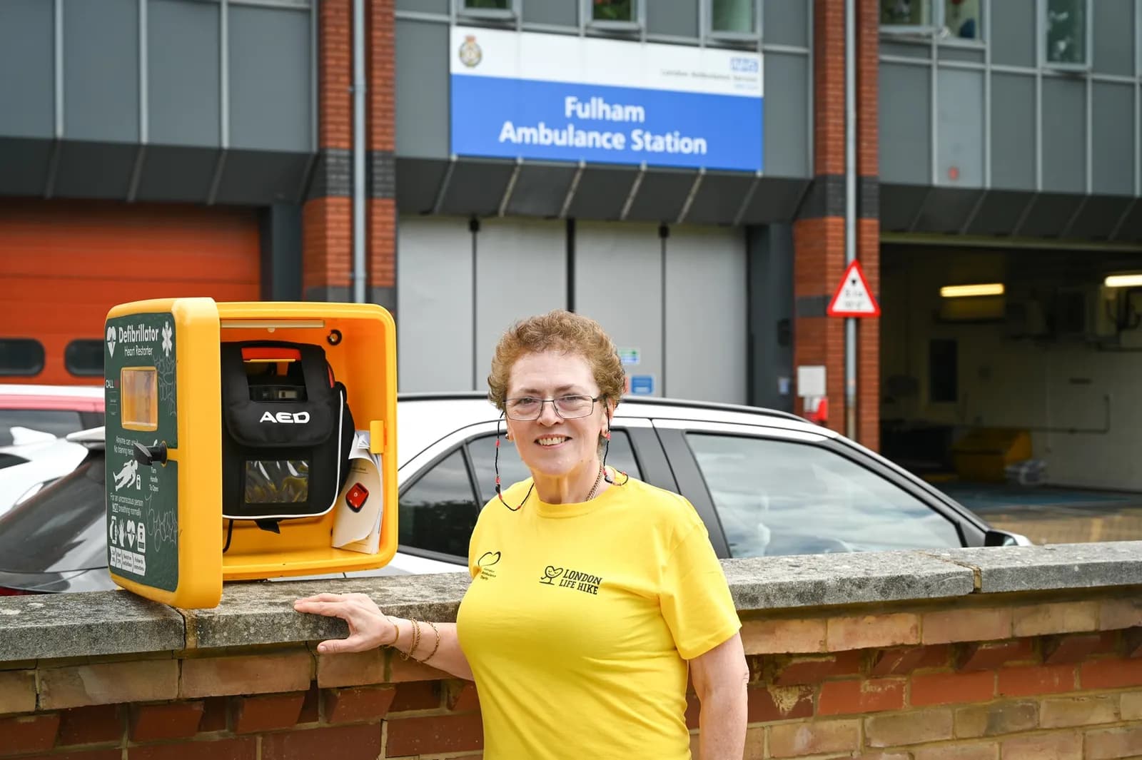 A smiling woman in a yellow shirt stands next to an open yellow defibrillator cabinet on a brick wall, with the Fulham Ambulance Station in the background.