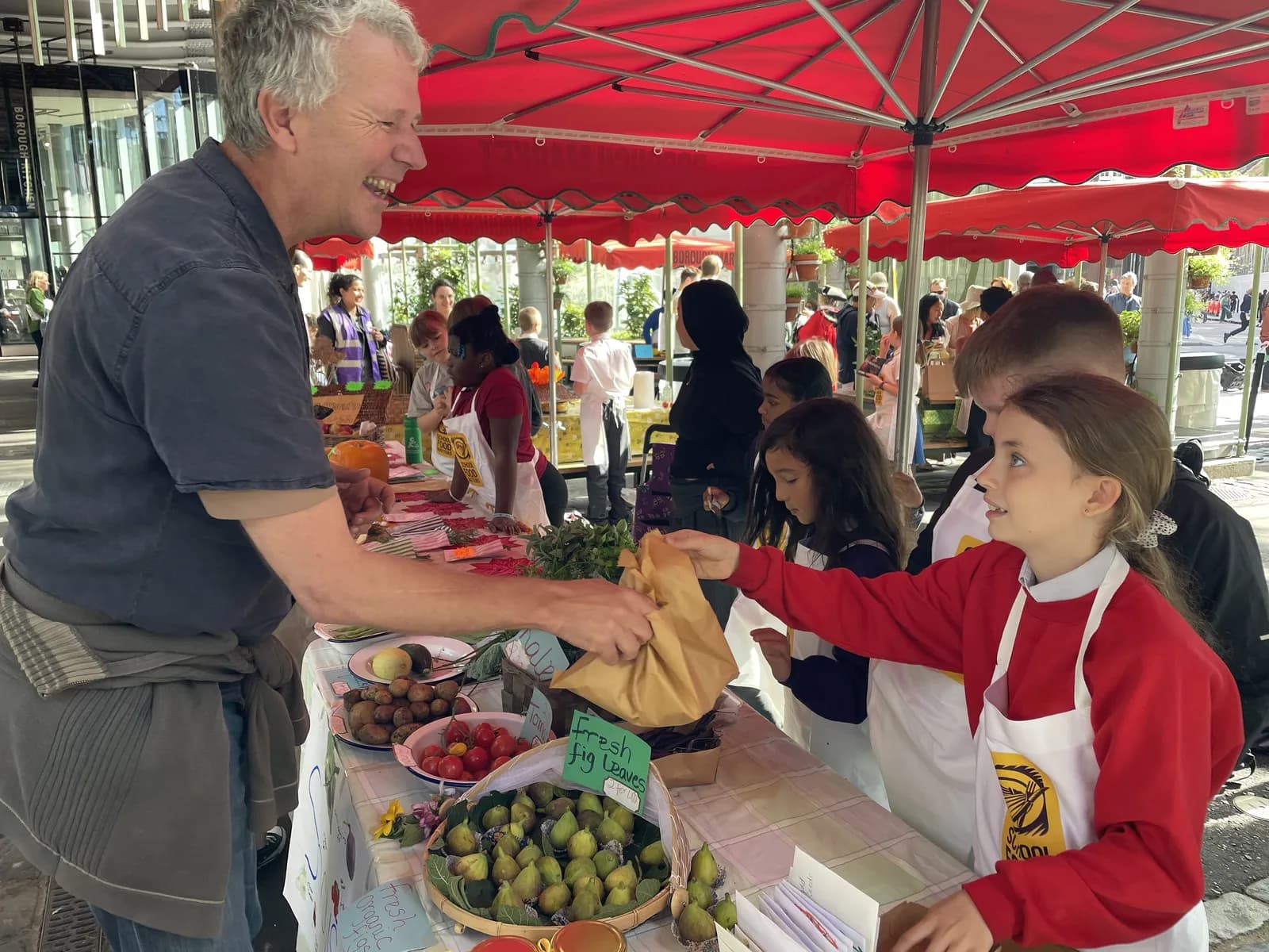 A man smiling as he hands a paper bag to a young girl at a market stall filled with fresh produce.