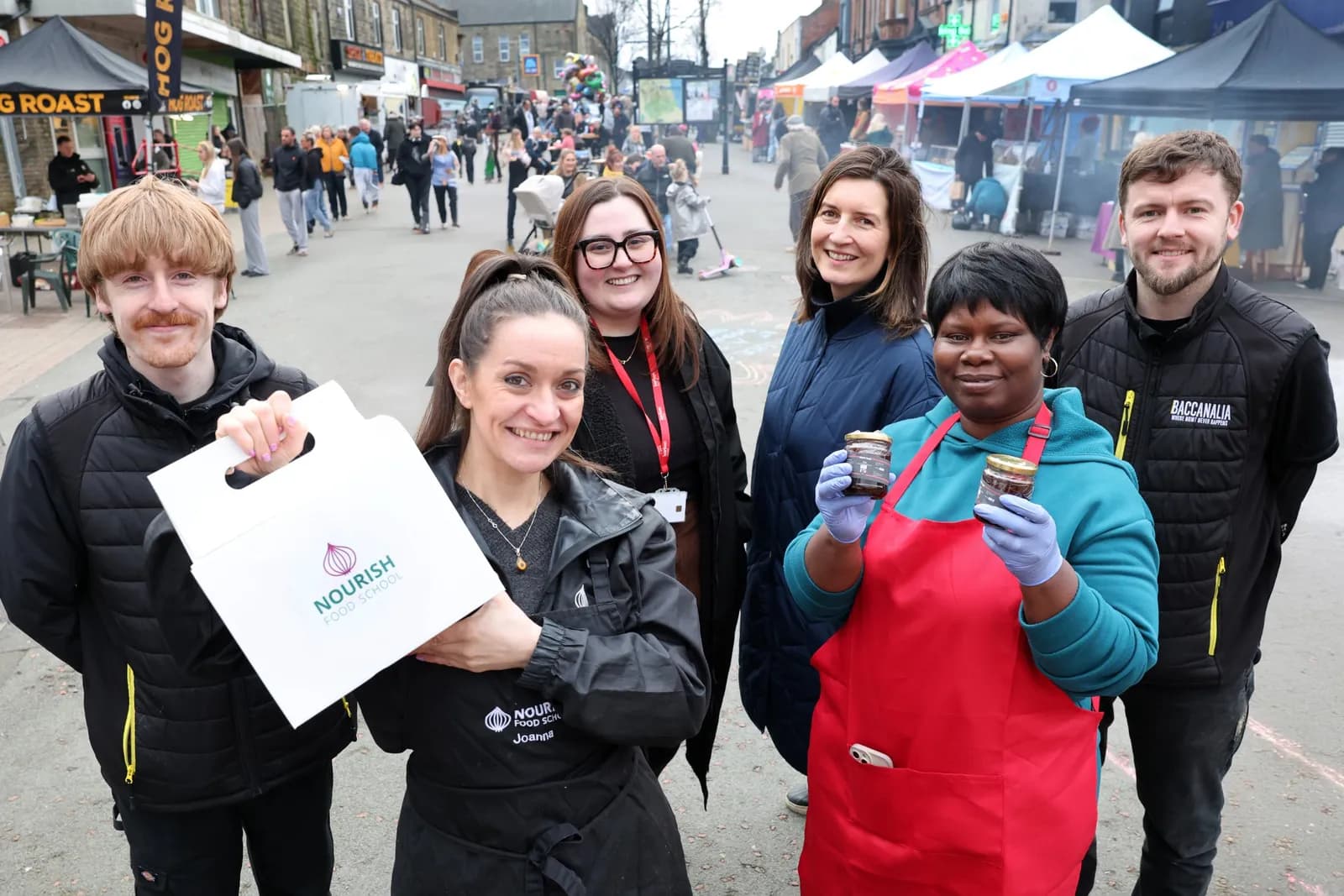 Six people, three women and three men, standing and smiling towards the camera, with a street fair in the background.