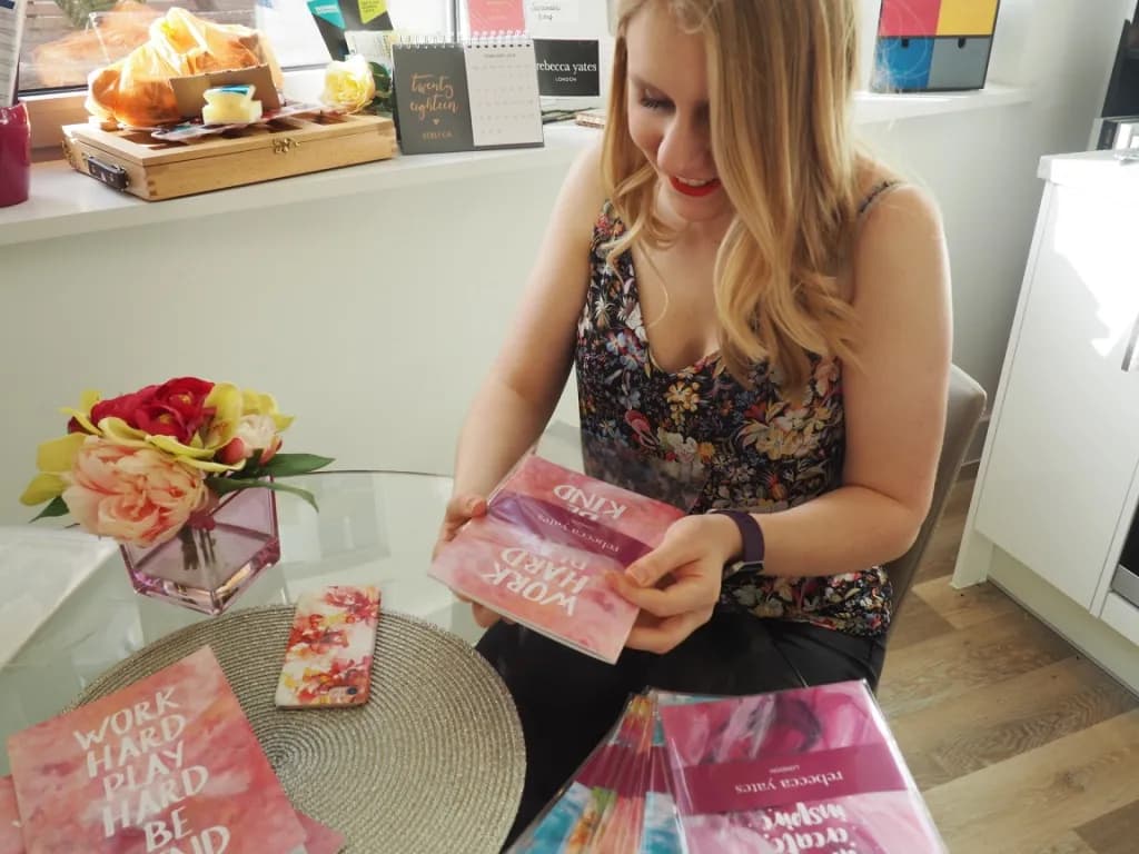A woman with blonde hair and a floral top is holding two books and smiling while sitting at a glass table with flowers and other books.