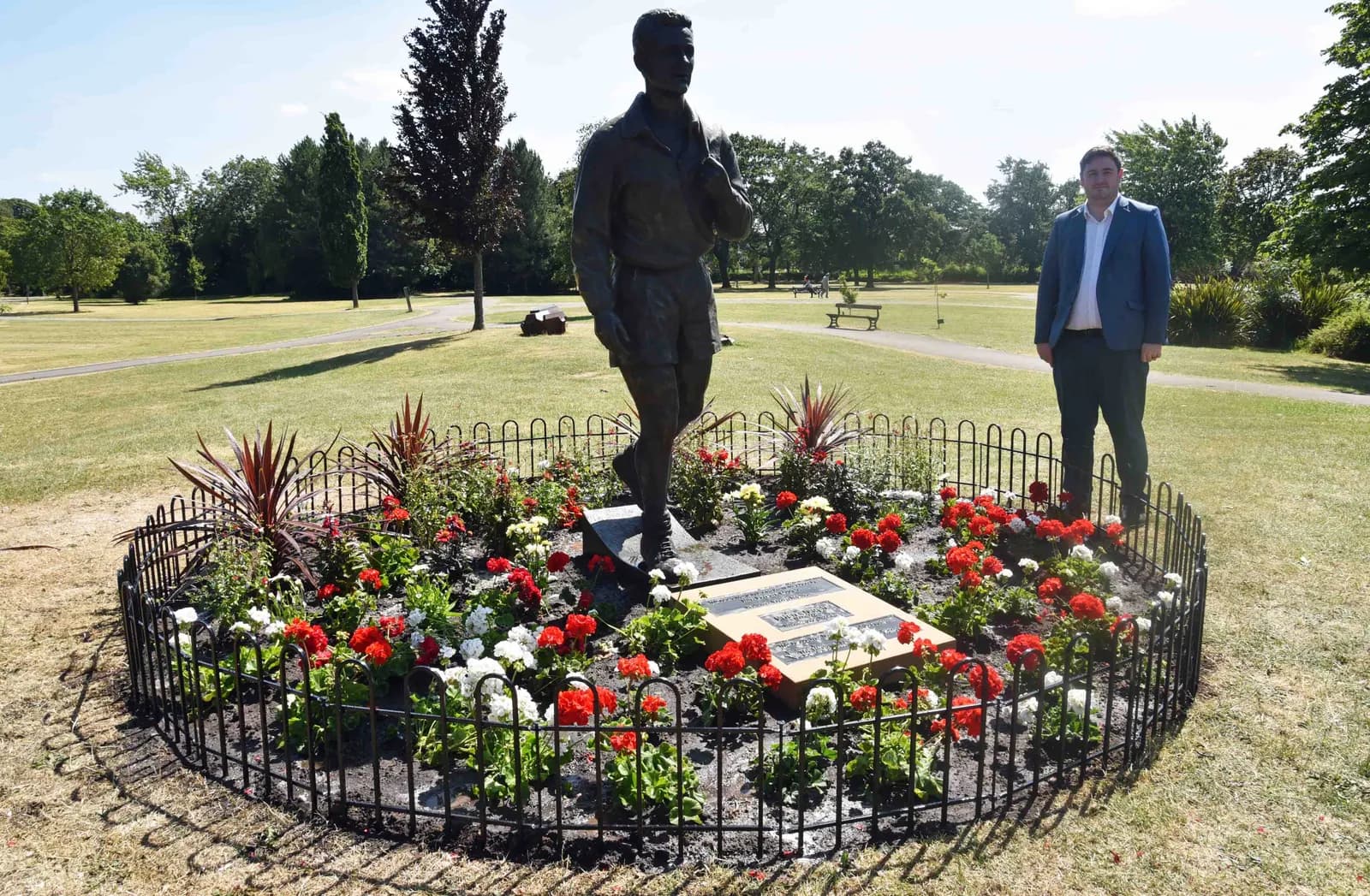 A man in a suit stands beside a bronze statue of a man in athletic wear, both positioned within a circular flower bed in a park.