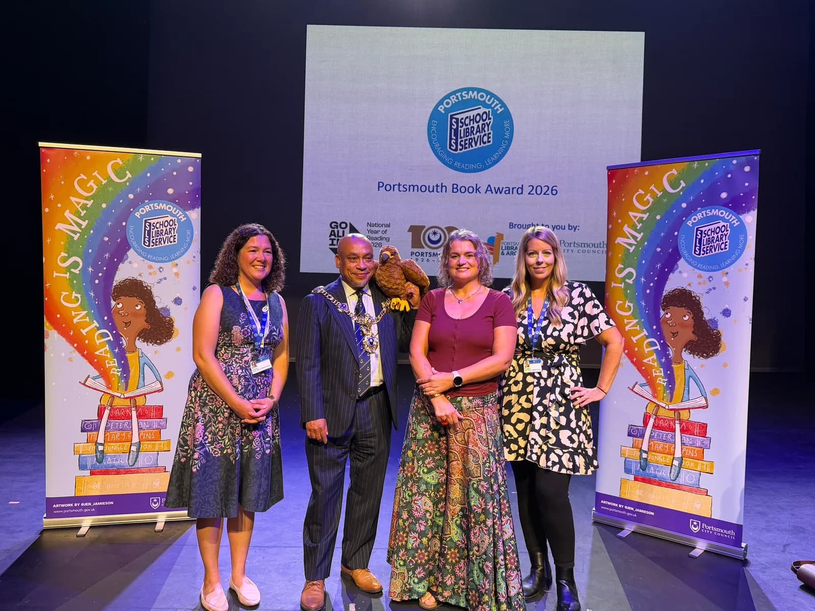 Four people, two men and two women, stand smiling in front of a stage backdrop for the Portsmouth Book Award 2026, flanked by colorful 'Reading is Magic' banners.
