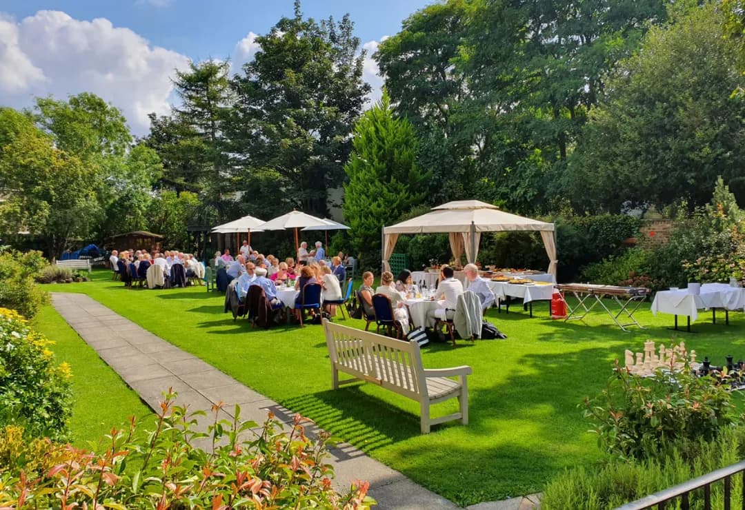 A large outdoor garden party with many people seated at round tables on a sunny green lawn, shadowed by trees, with food tables and white umbrellas and canopies.