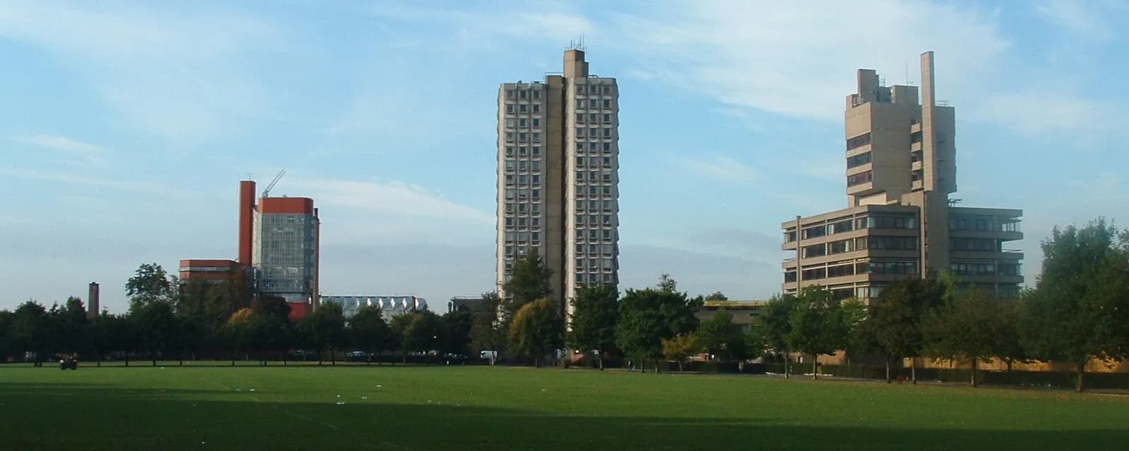 A wide shot shows three tall, distinct architectural buildings standing against a light blue sky, two on the right and one on the left, with a green grassy field and a line of trees in the foreground.