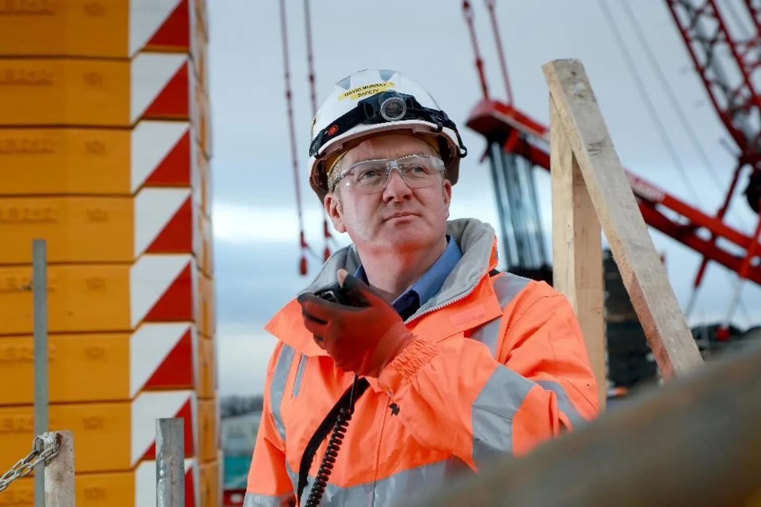 A man in an orange safety jacket, hard hat, and safety glasses holds a two-way radio at a construction site.