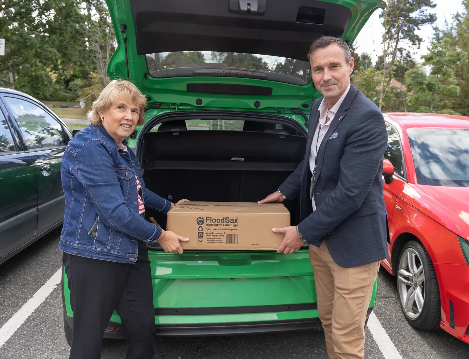 A woman in a denim jacket and a man in a suit are placing a box labeled "FloodSax" into the open trunk of a green car.