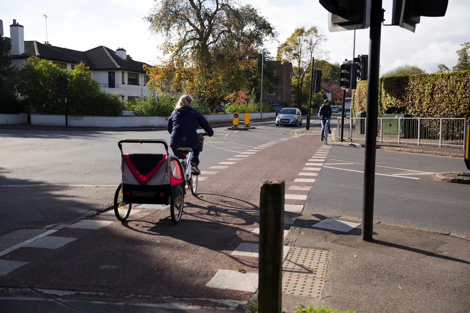 A woman on a bicycle with a child trailer attached crosses a street, with another cyclist and a car visible in the background.