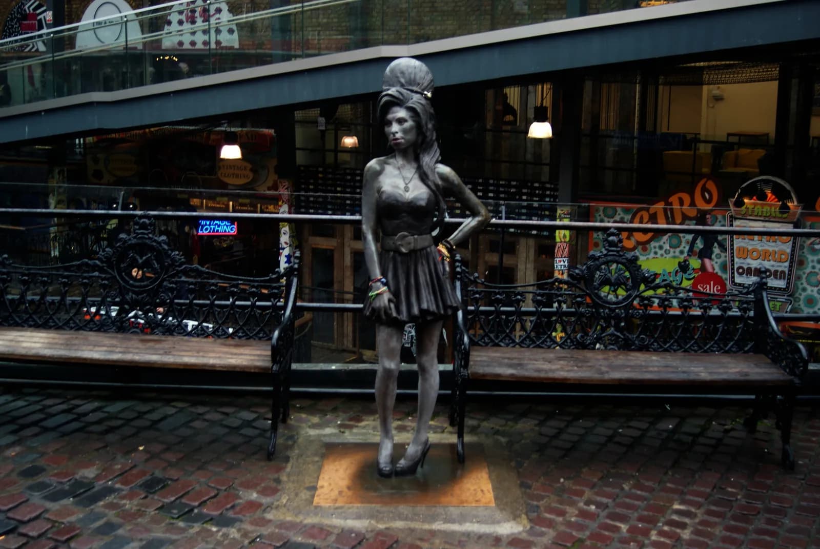 A life-sized bronze statue of Amy Winehouse stands between two benches on a brick path, with building storefronts and an upper-level walkway in the background.
