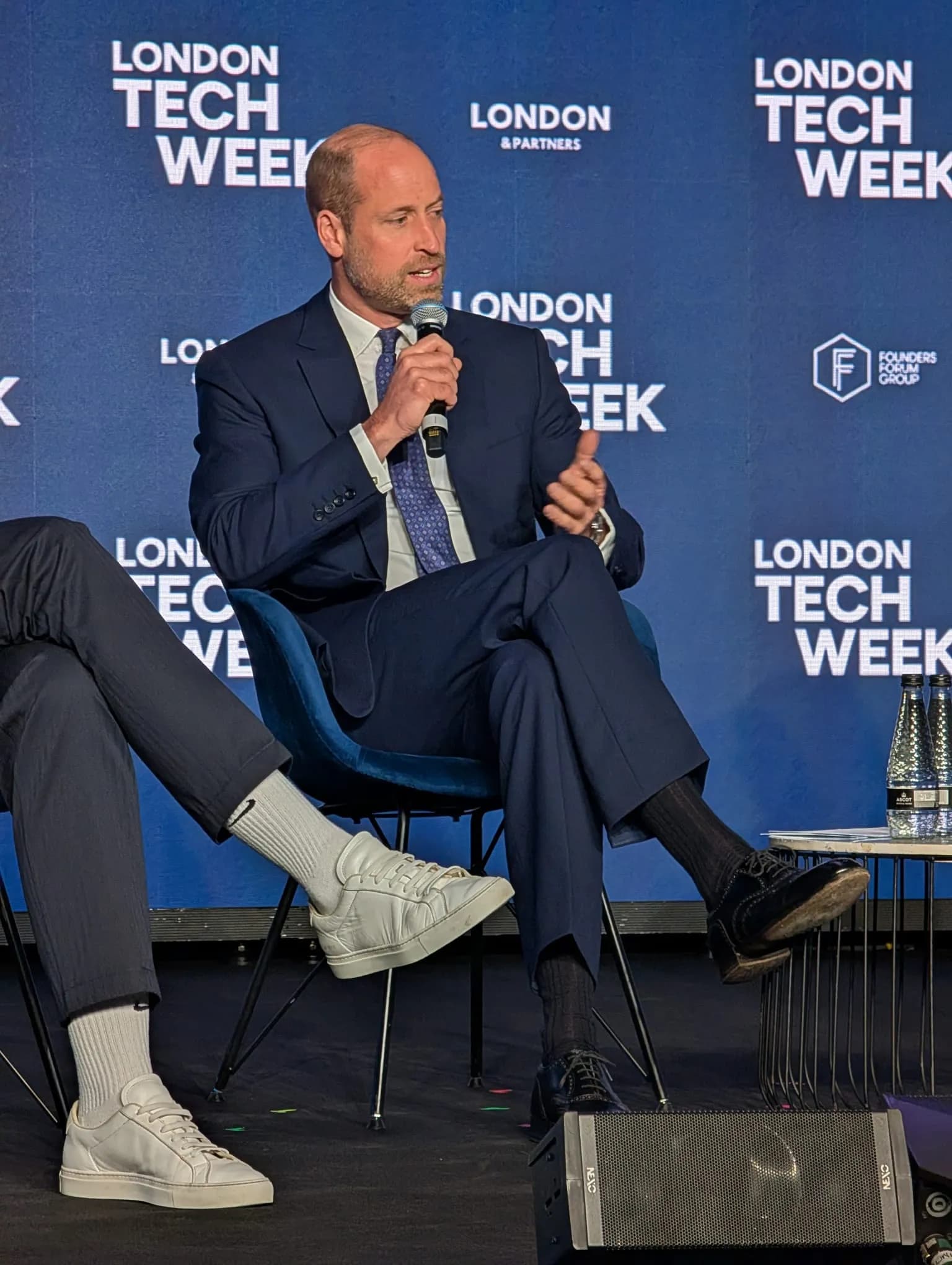 Prince William speaking on stage at London Tech Week, dressed in a blue suit and holding a microphone.