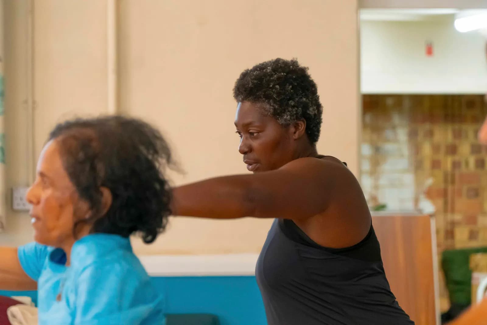 A woman with short, curly, graying hair in a black tank top extends her arm forward during an exercise class, with another person partially visible in the foreground.