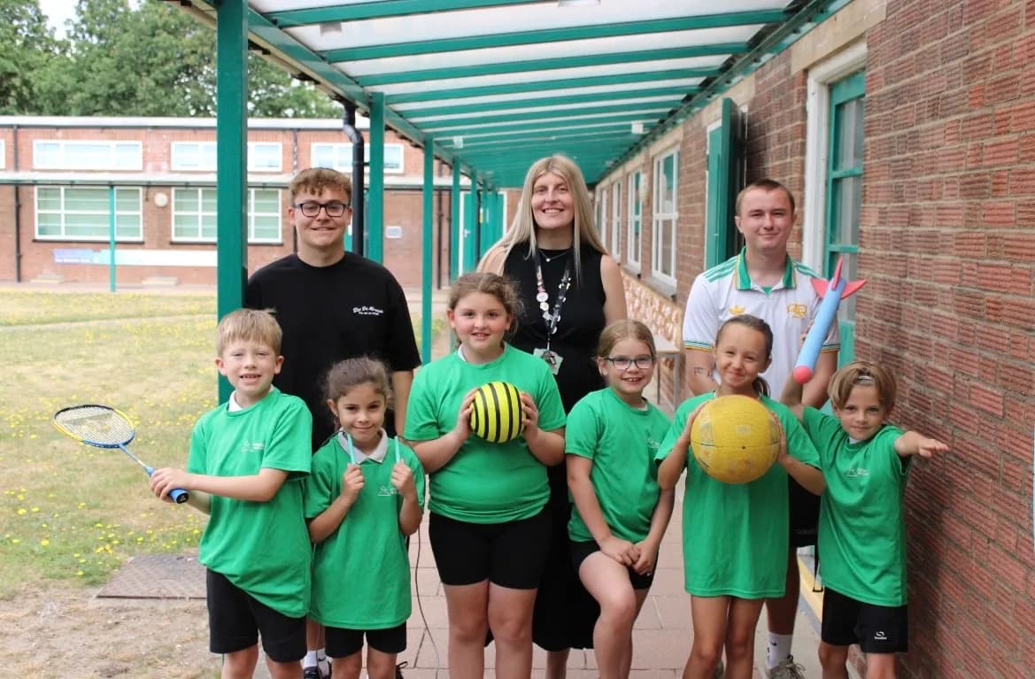 A group of children and adults, all dressed in green shirts, stand in front of a school building while holding various sports equipment.