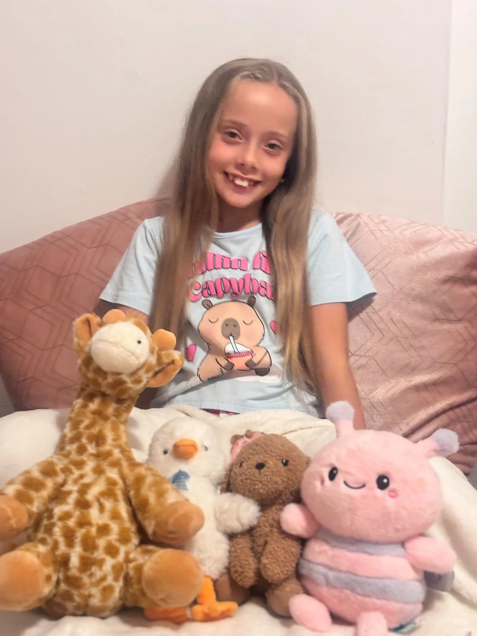 A smiling girl with long brown hair sits on a bed with a pink pillow behind her, surrounded by a giraffe, duck, bear, and pink bee stuffed animals.