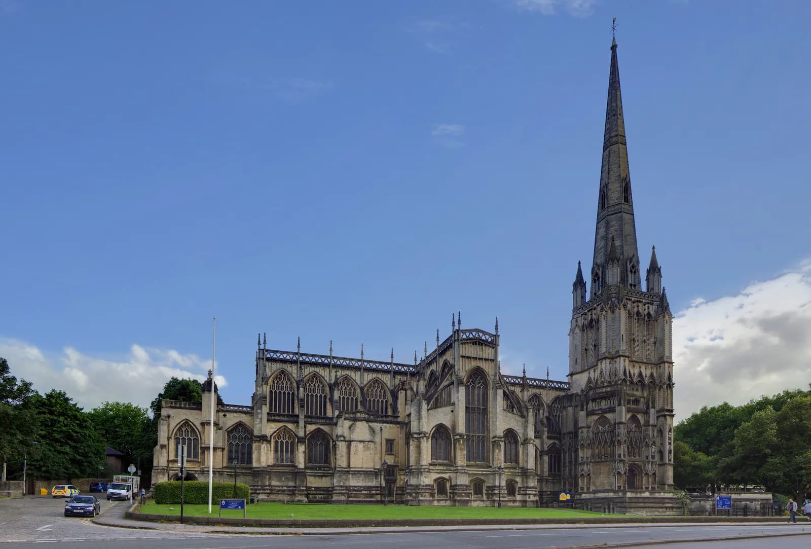 A grand, old cathedral with a tall, slender spire stands beside a road with a small patch of grass and trees under a bright blue sky with some clouds.