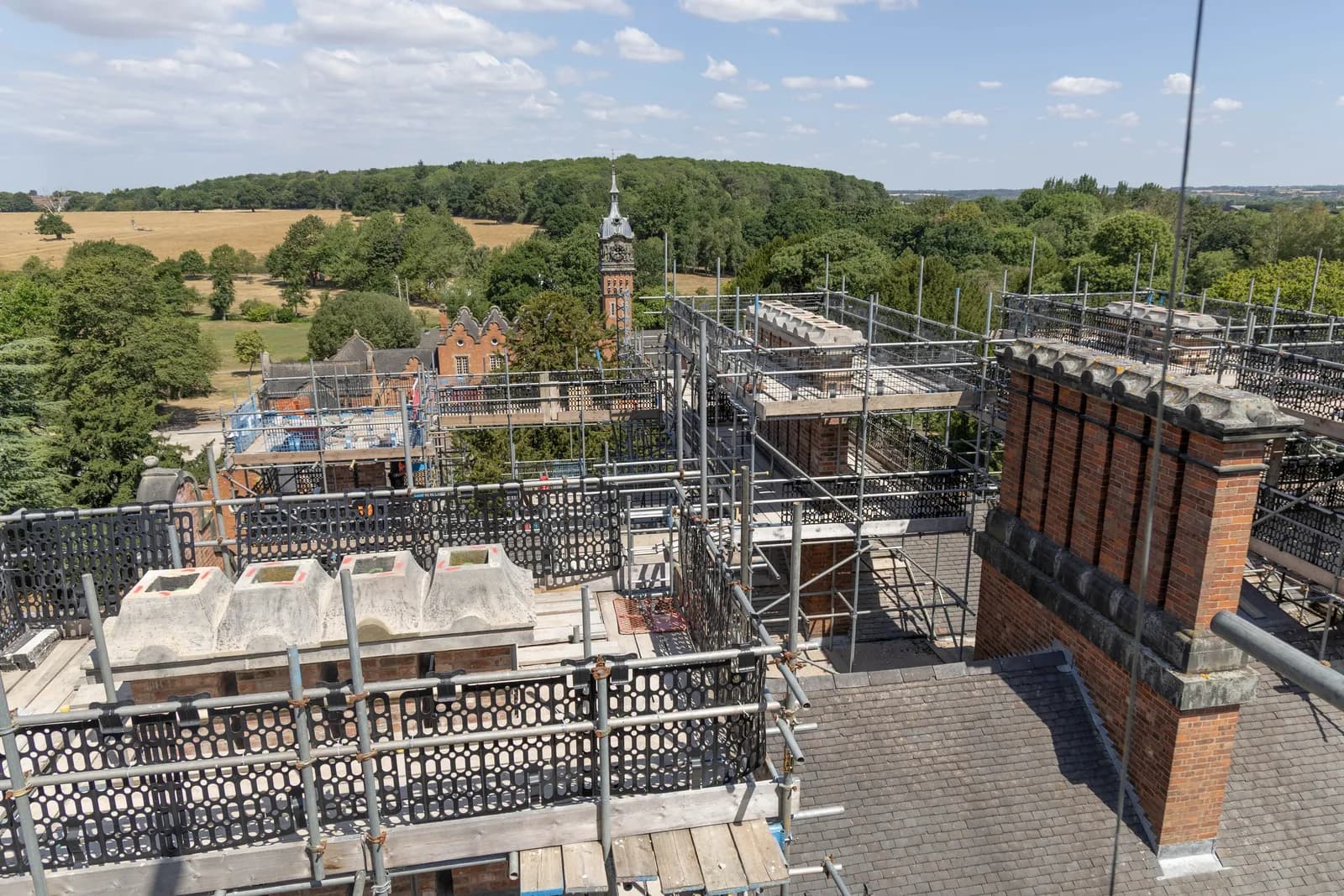 A high-angle view shows a large brick building with scaffolding around its roof and chimneys, surrounded by trees and a distant field under a cloudy sky.