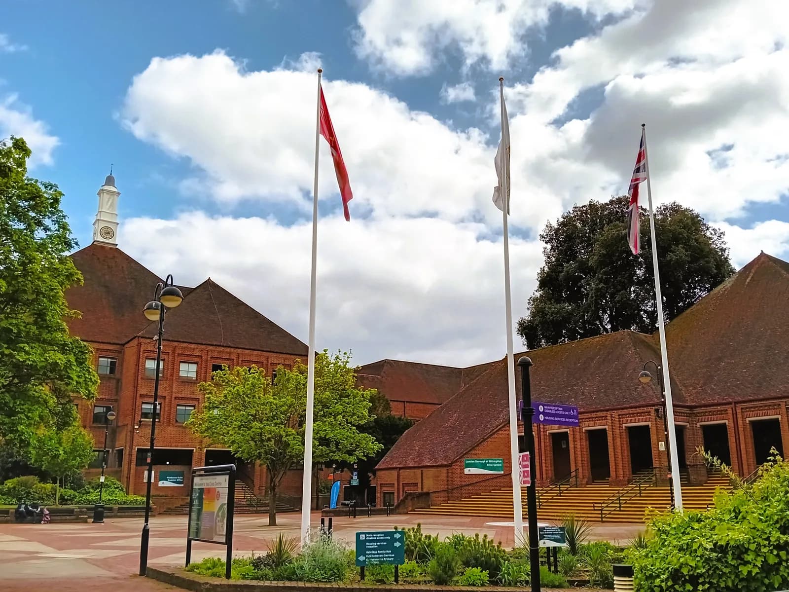A red brick building with a white clock tower on the left, three white flagpoles with a red, white, and Union Jack flag, and another brick building on the right under a blue sky with white clouds.