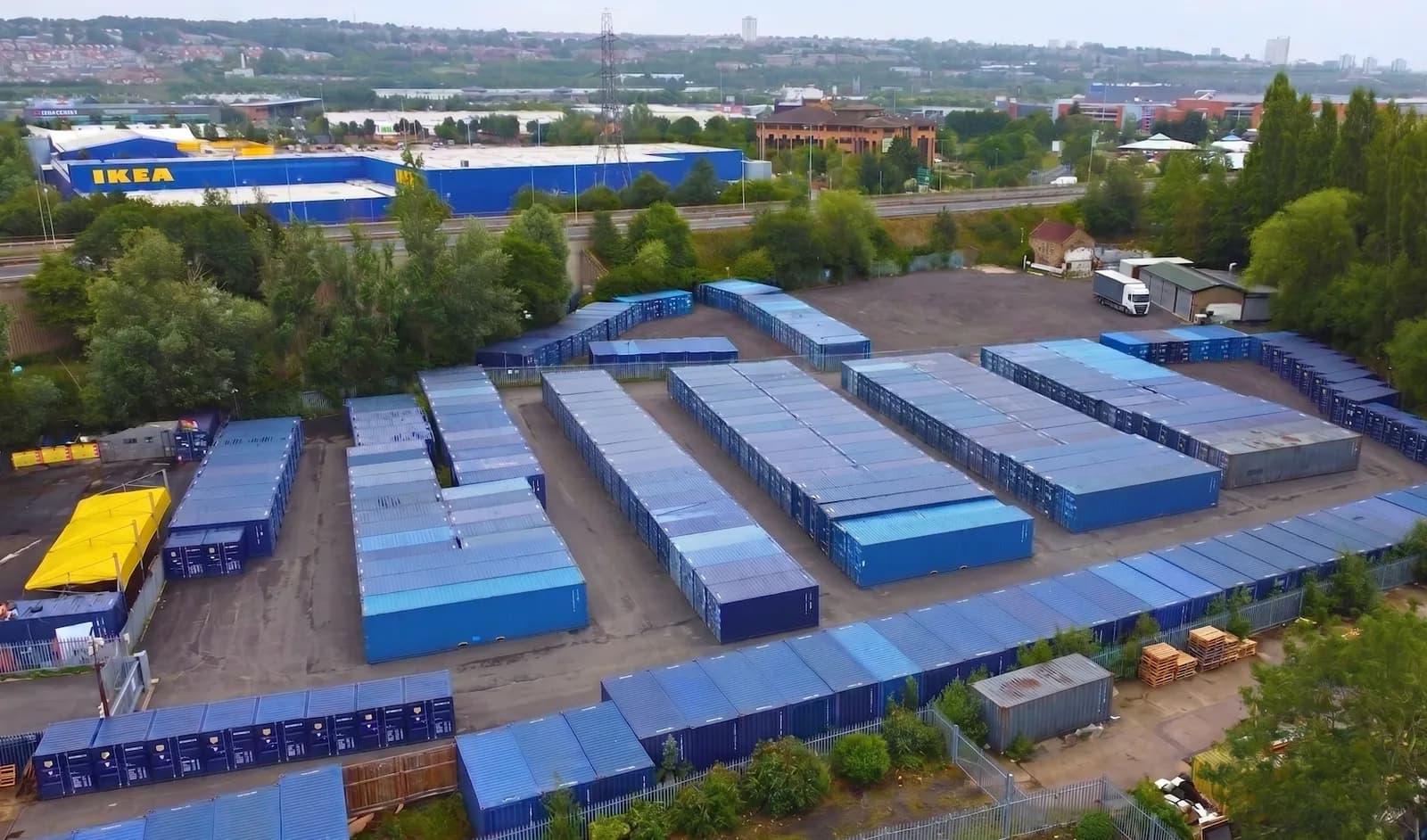 An aerial view shows a large outdoor storage facility with rows of blue shipping containers, some covered with tarps, and an IKEA store in the background.