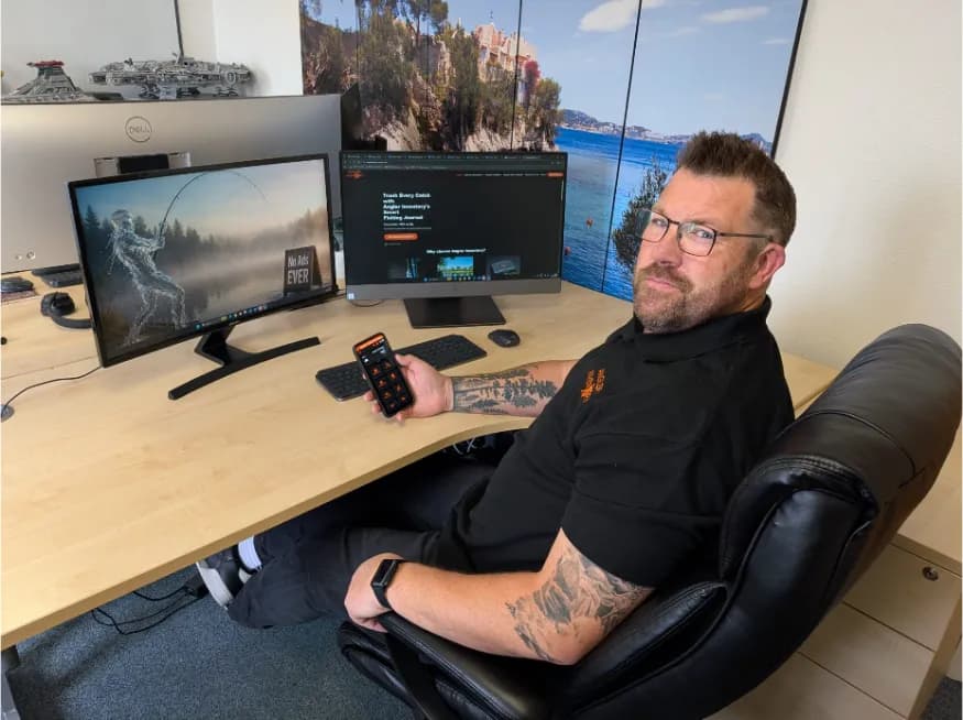 A man with glasses and a beard sits at a desk with two computer monitors, holding a smartphone in his right hand.